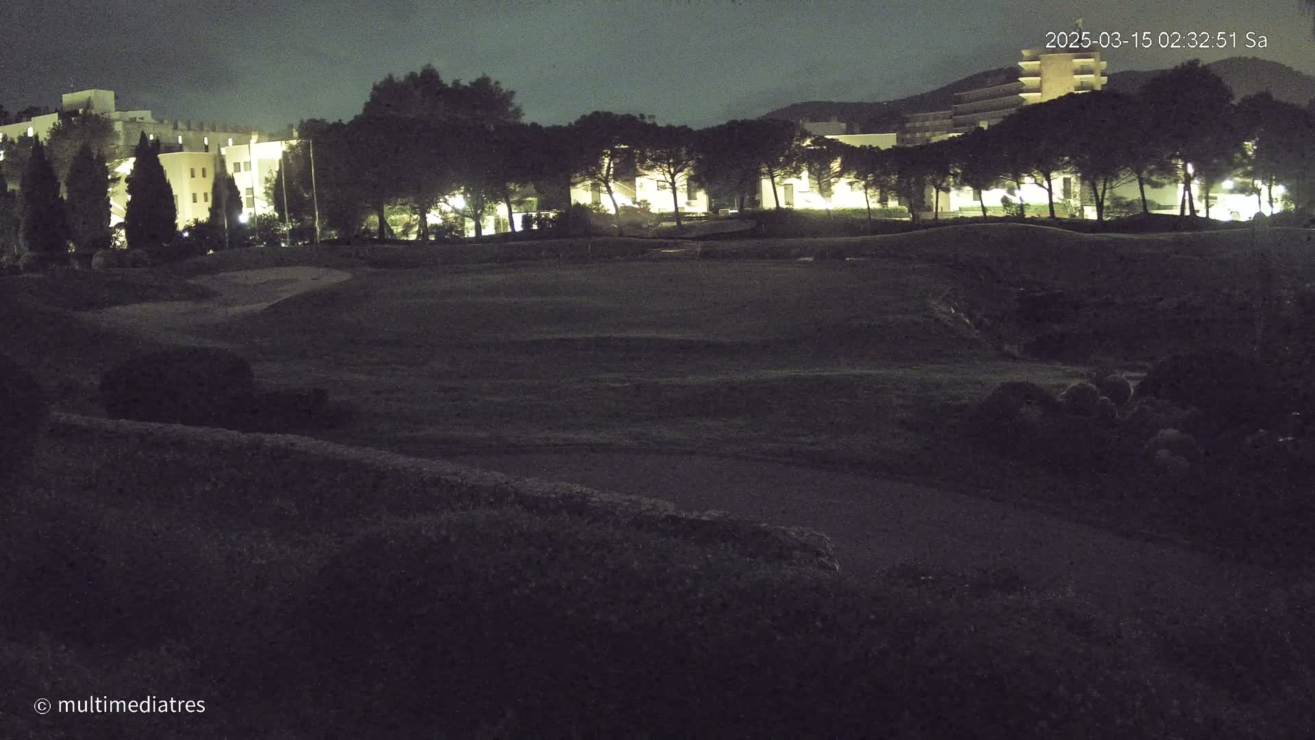 A nighttime view of a grassy area with illuminated buildings and trees in the background.