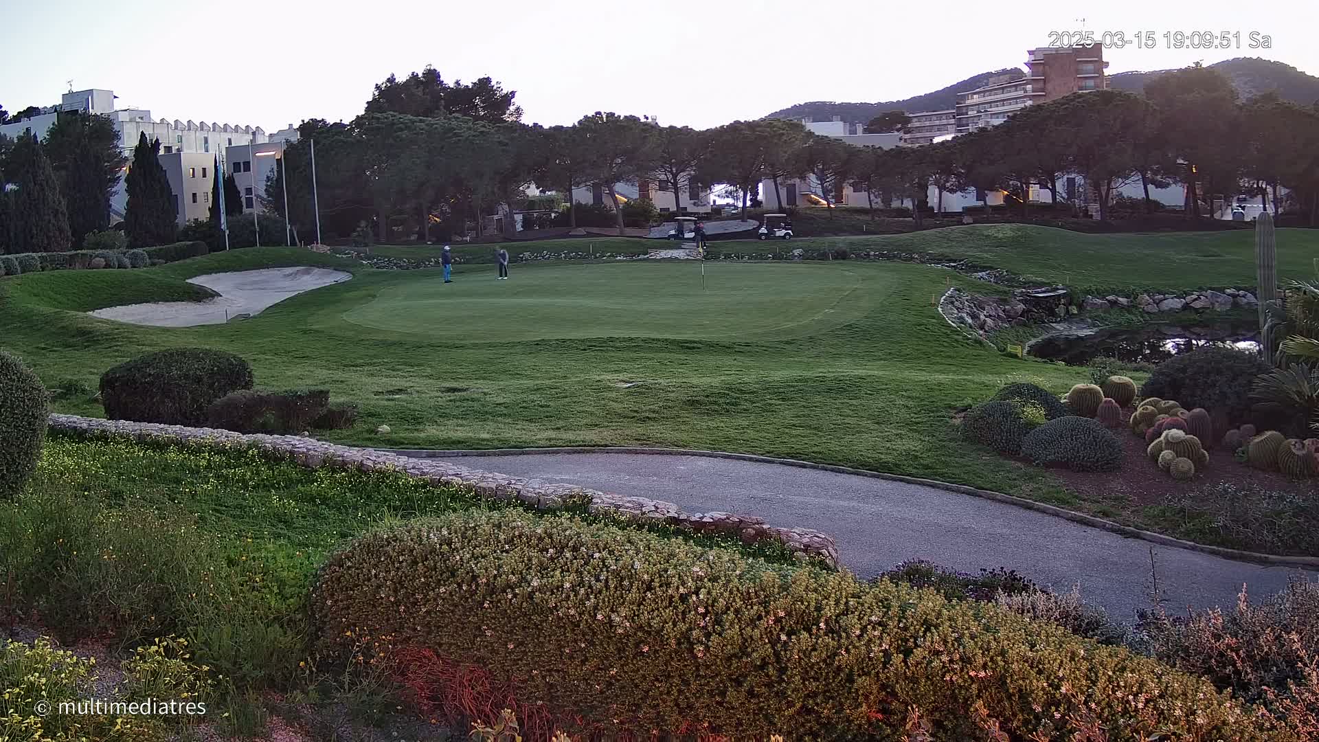 A golf course green with several golfers and golf carts is visible, with a sand trap and landscaping features including cacti, under a clear twilight sky.