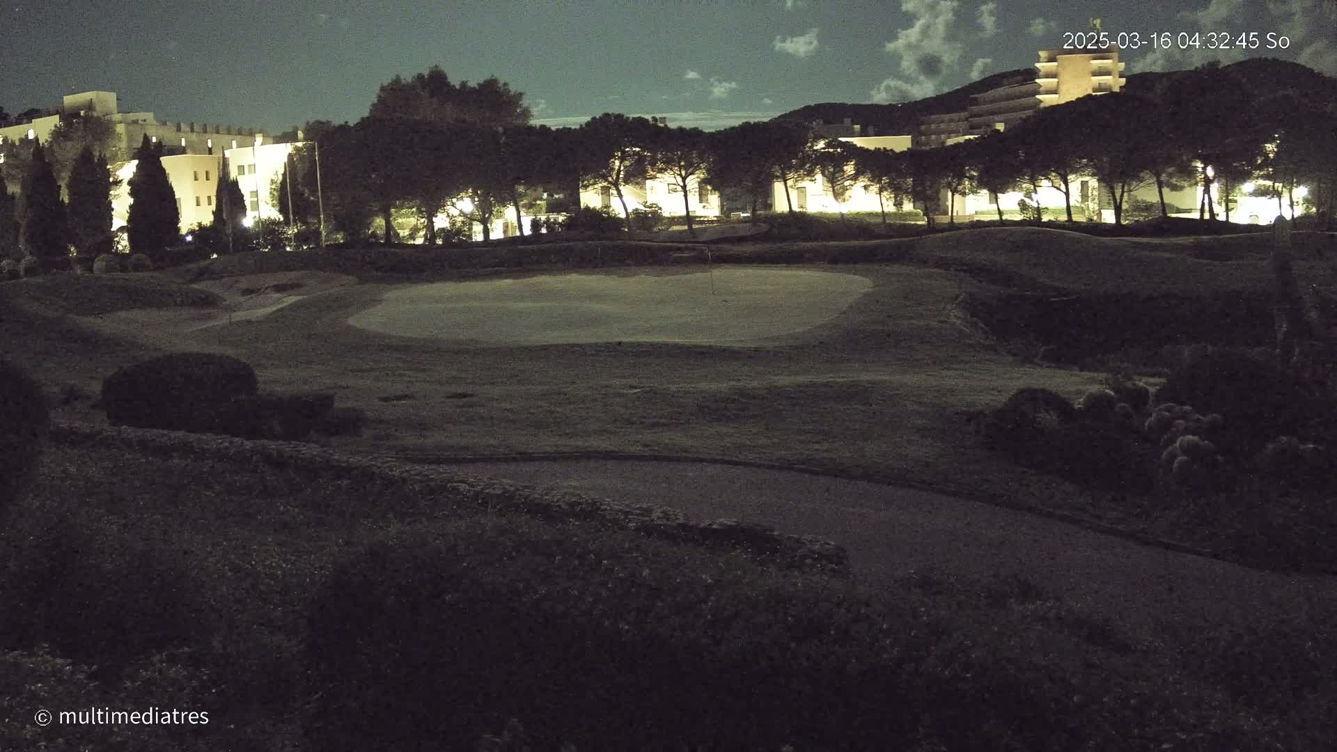 A nighttime view of a golf course with a building and trees in the background under a slightly cloudy sky.