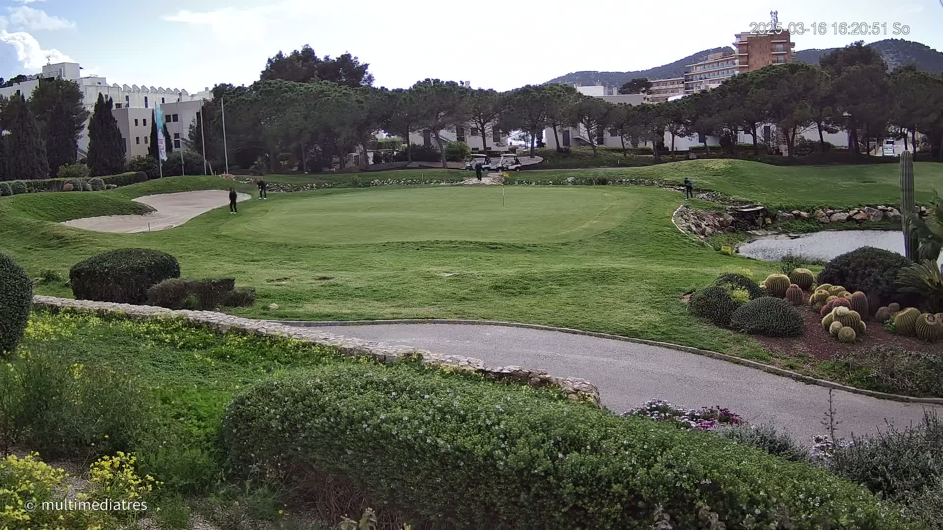 A golf course green with several golfers, bordered by landscaping, a small pond, and buildings under a partly cloudy sky.
