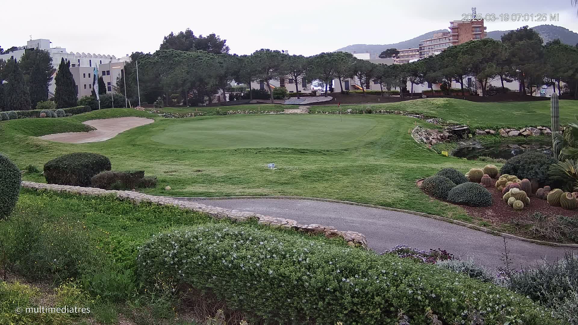 A golf course putting green is shown, with a sand trap, landscaping, and buildings in the background under an overcast sky.