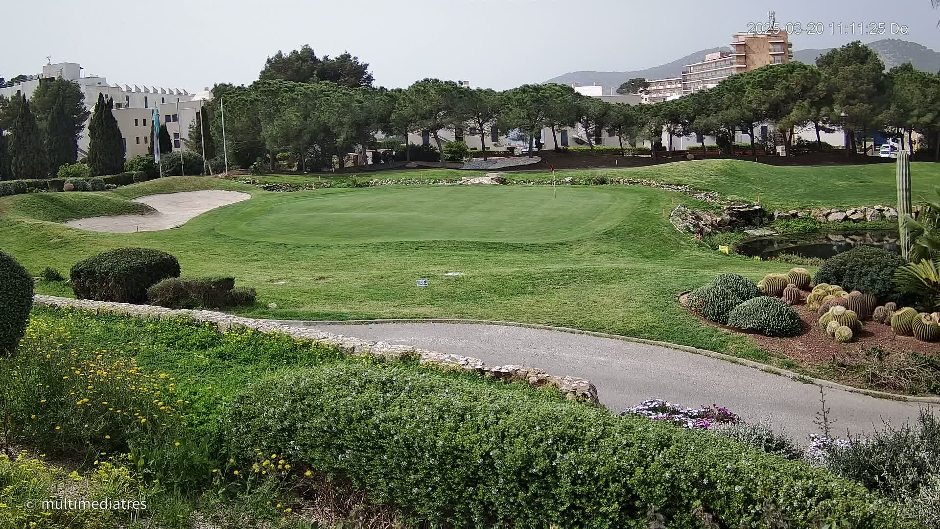 A golf course green with sand traps, a small pond, and cacti is visible in the foreground, with a hotel and other buildings in the background under a partly cloudy sky.