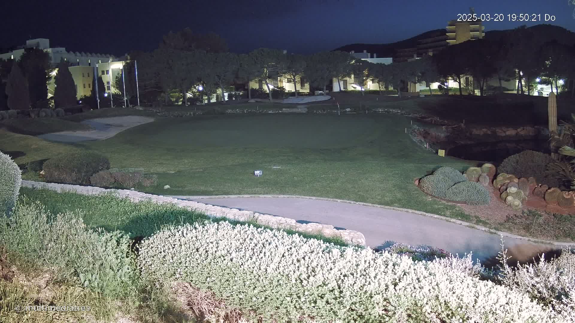 A nighttime view of a putting green next to a building complex, illuminated by artificial lights, with low shrubs in the foreground.