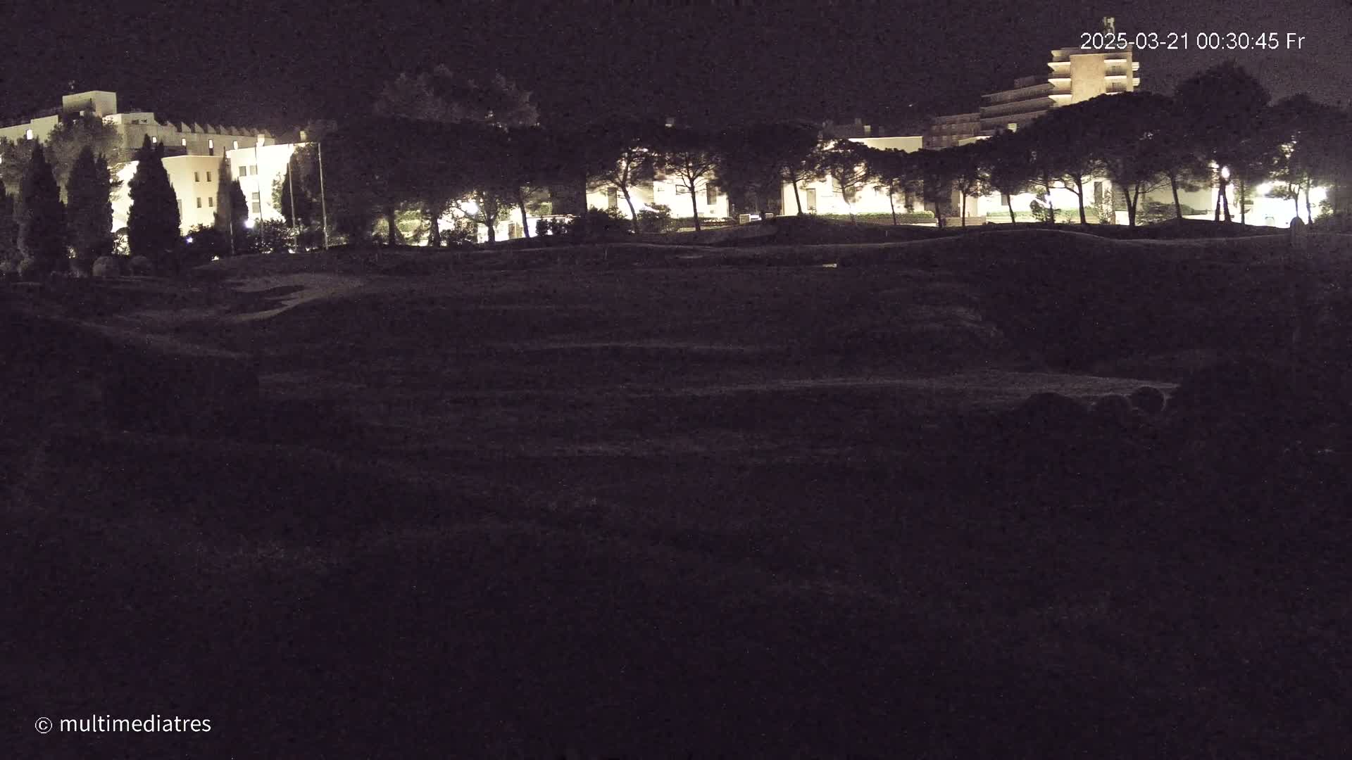 A nighttime view shows a lighted building behind a row of dark trees and a large, dark, uneven field.