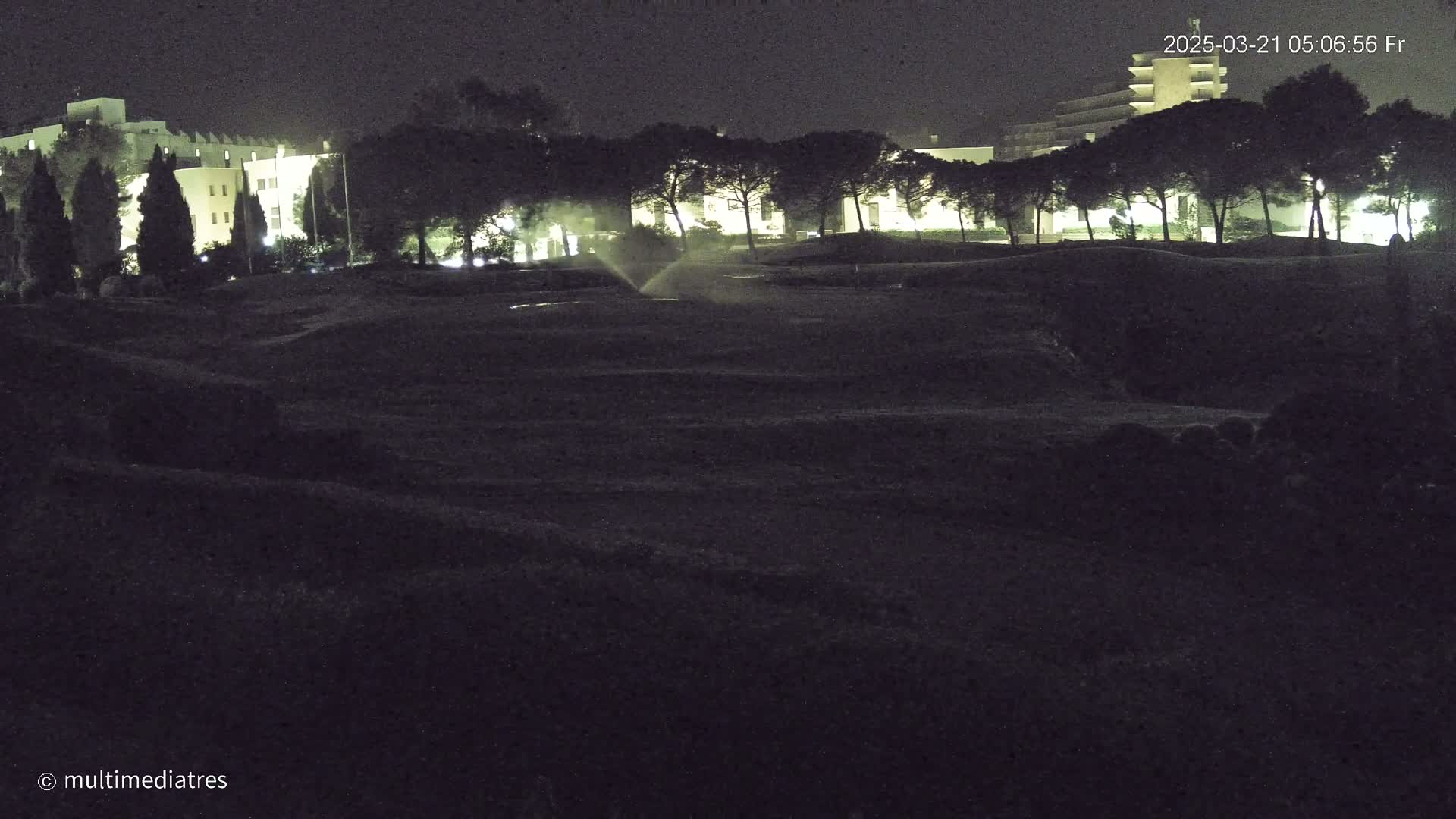 A nighttime view of a grassy area with illuminated buildings and trees in the background, under dark skies.