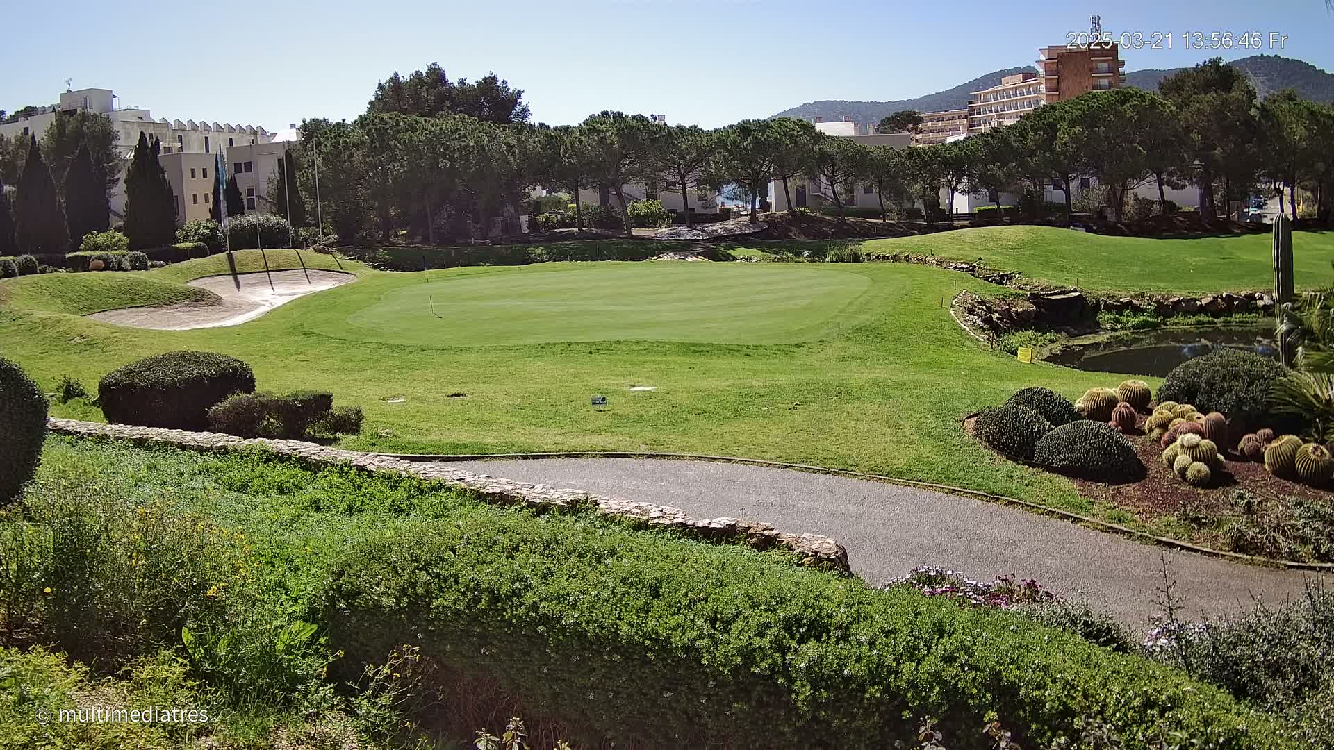 A golf course green with a sand trap and landscaped areas, under a clear sunny sky, is situated near buildings and trees.