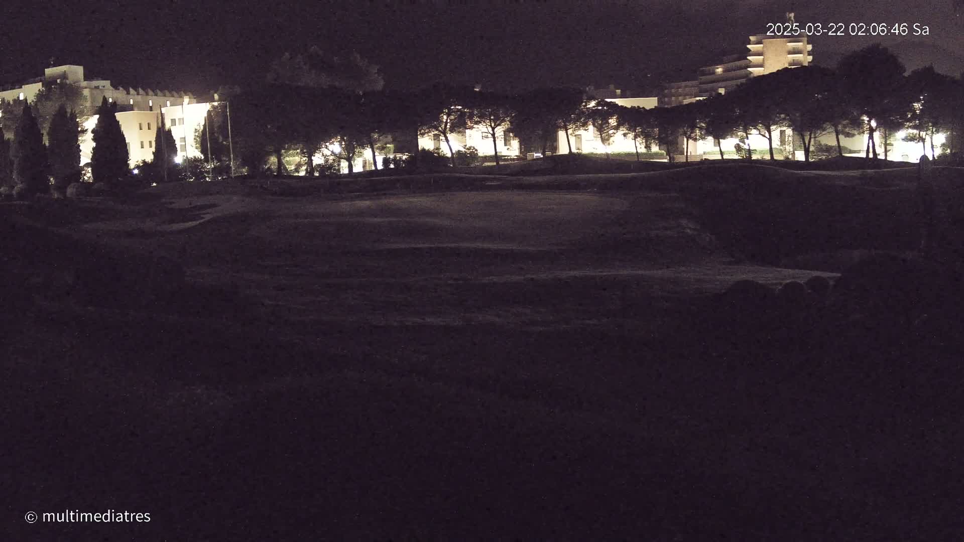At night, a dark, grassy expanse stretches before illuminated buildings and a row of trees.