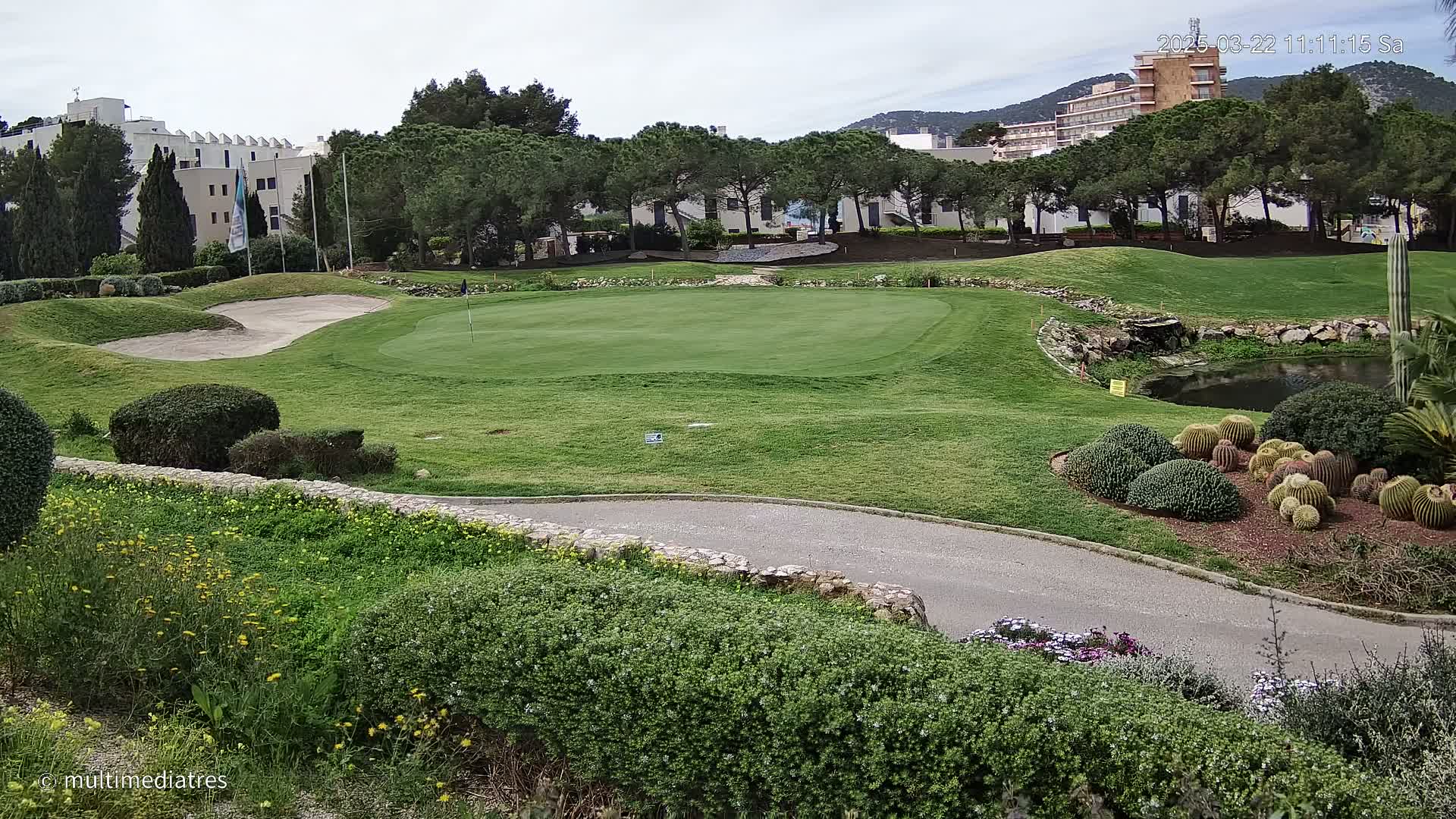 A golf course green with a sand trap, bordered by low hedges and cacti, is situated near buildings under a partly cloudy sky.