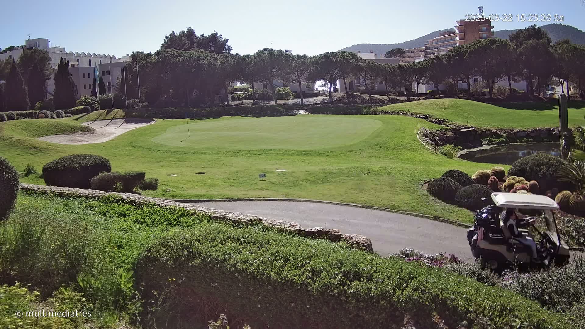 A golf course green with a golf cart on a paved path, surrounded by landscaping and buildings in the background under sunny skies.