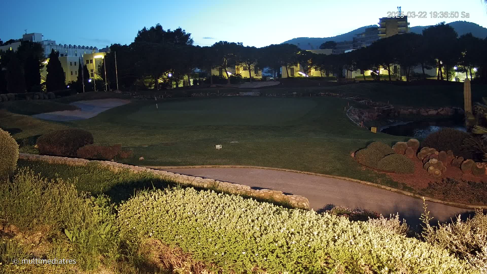 A well-lit putting green and surrounding landscaping at twilight, with buildings and trees in the background.