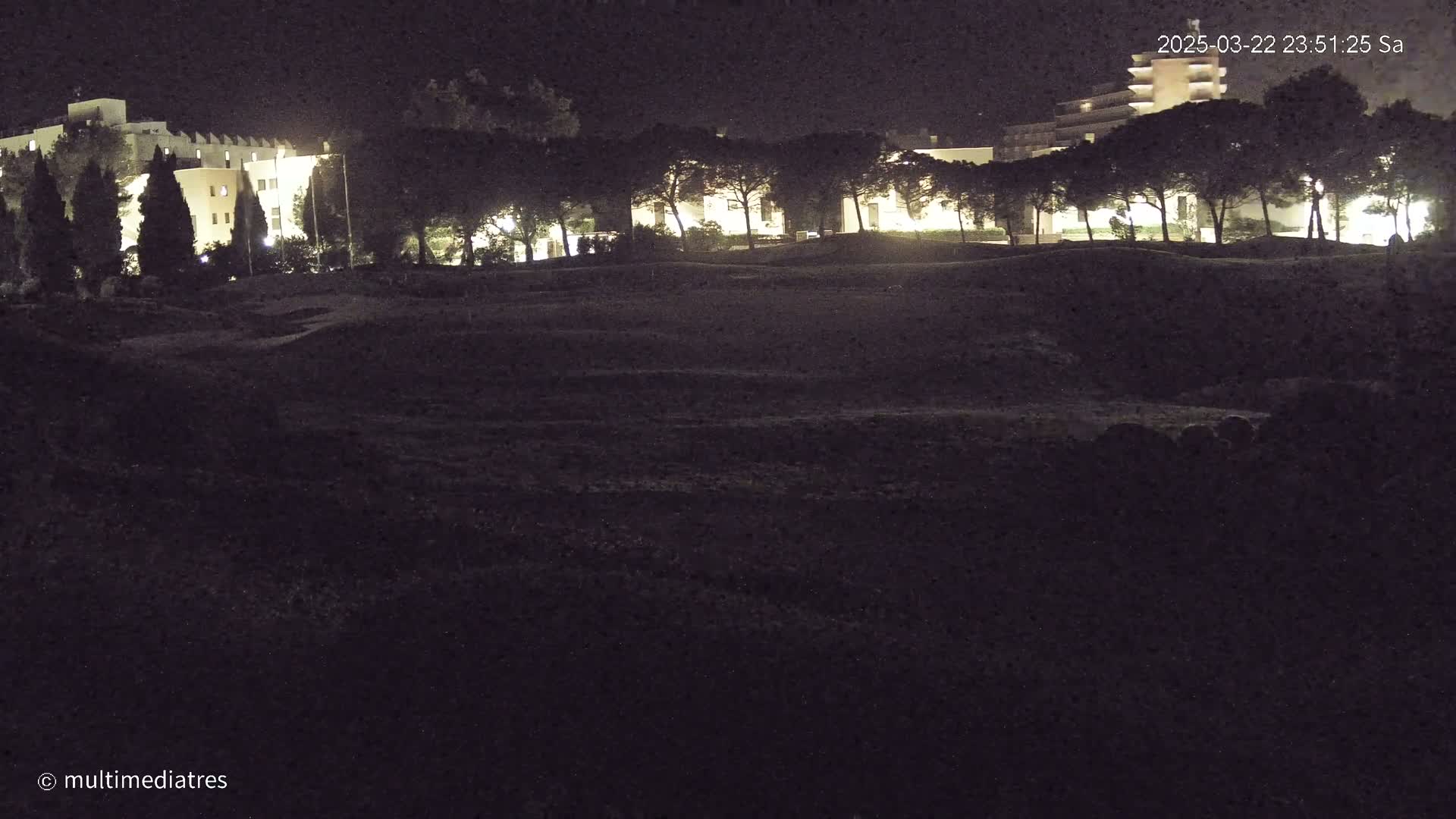 At night, a line of illuminated buildings is visible behind a row of dark trees, and a dark, uneven field stretches out in front.