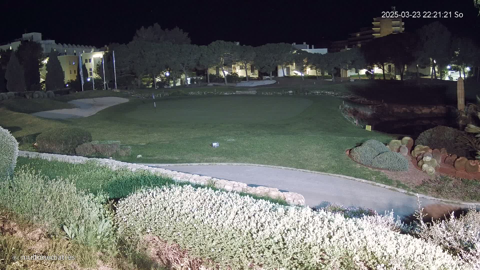A nighttime view of a putting green on a golf course, adjacent to buildings and landscaping, under artificial lighting.