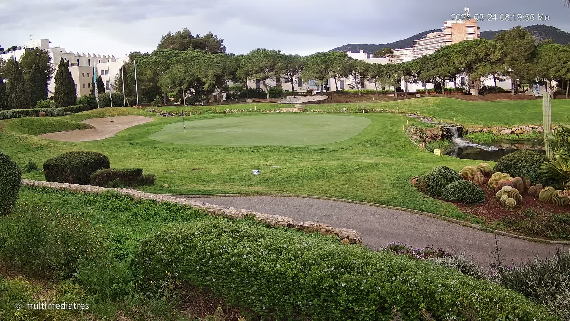A golf course green with a sand trap, a small pond, and a low stone wall, under a partly cloudy sky.