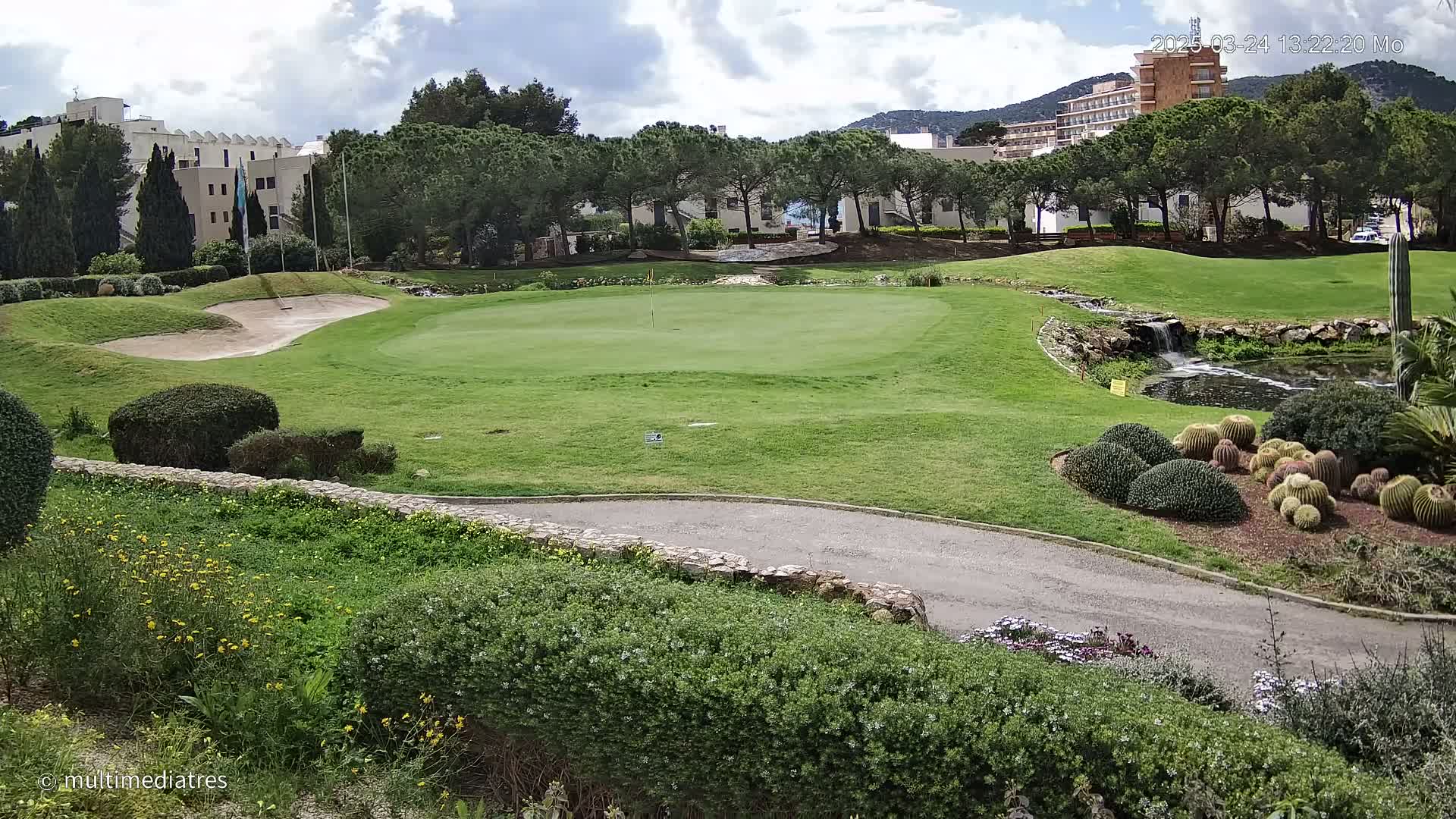 A golf course green with sand traps and a small pond, surrounded by lush landscaping and buildings under a partly cloudy sky.