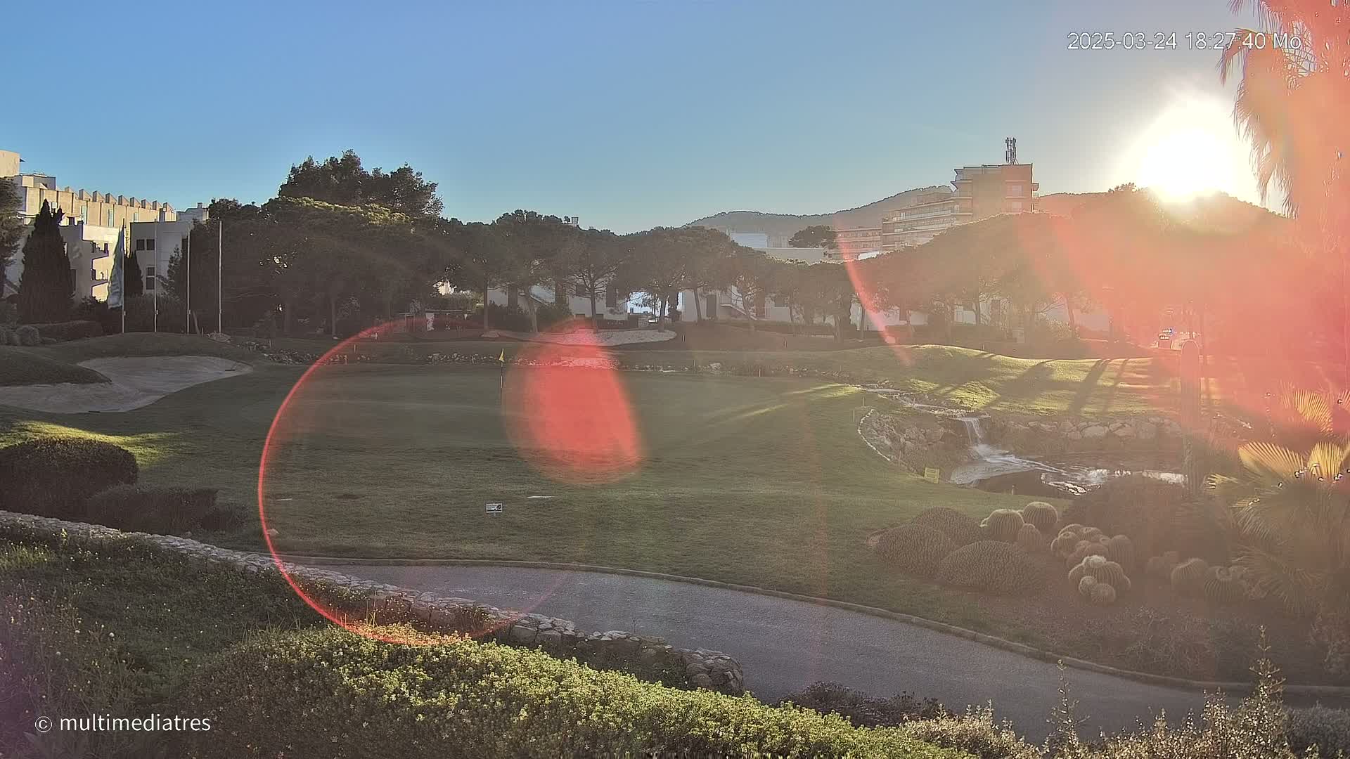 A sun-drenched golf course with a small waterfall and cacti garden, situated near buildings, under a clear, bright sky.