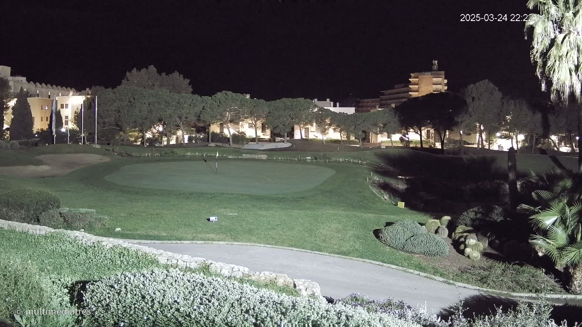 A lighted golf course green at night, with buildings and trees in the background.
