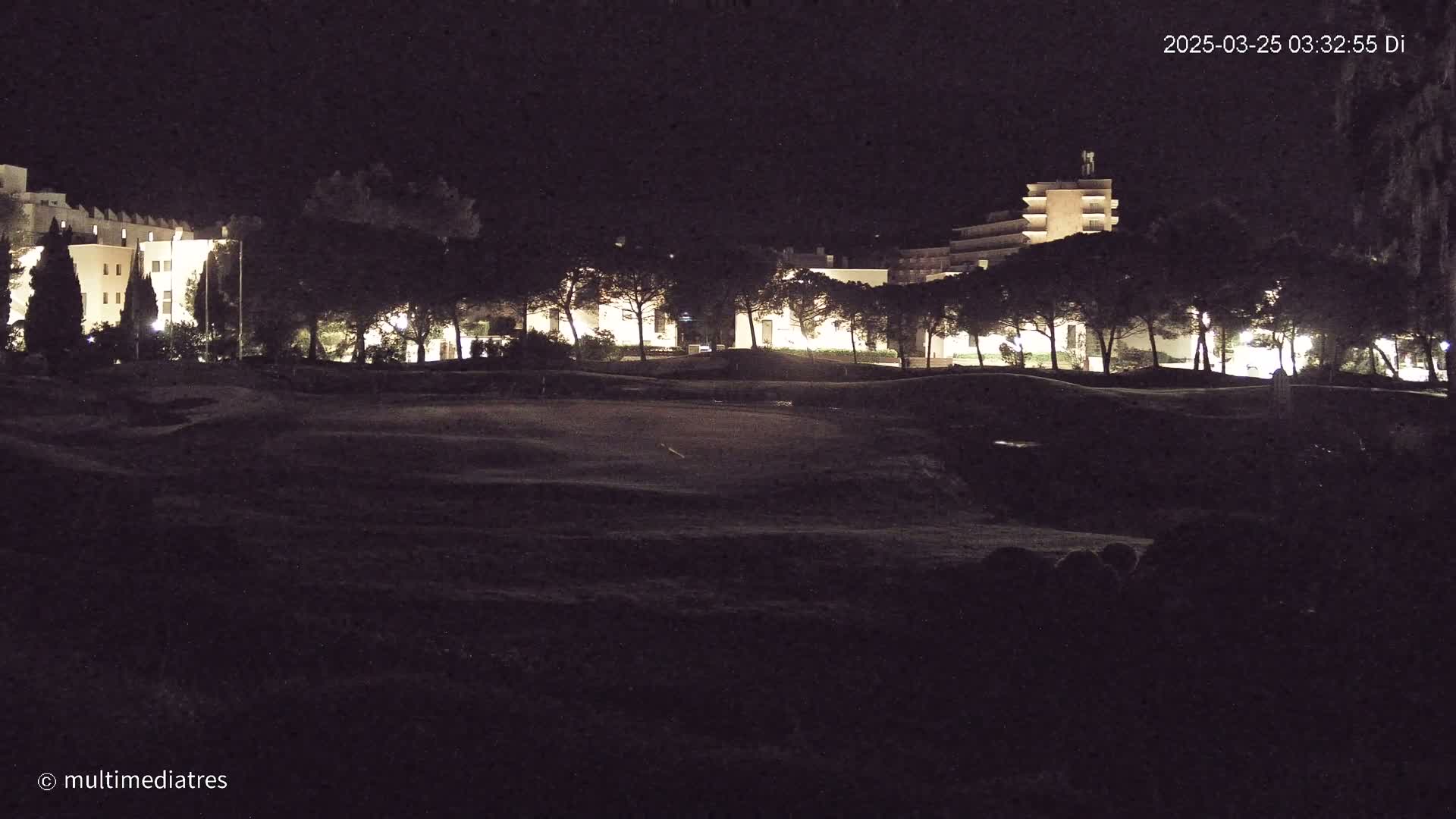 At night, illuminated buildings are visible beyond a dark, grassy expanse.