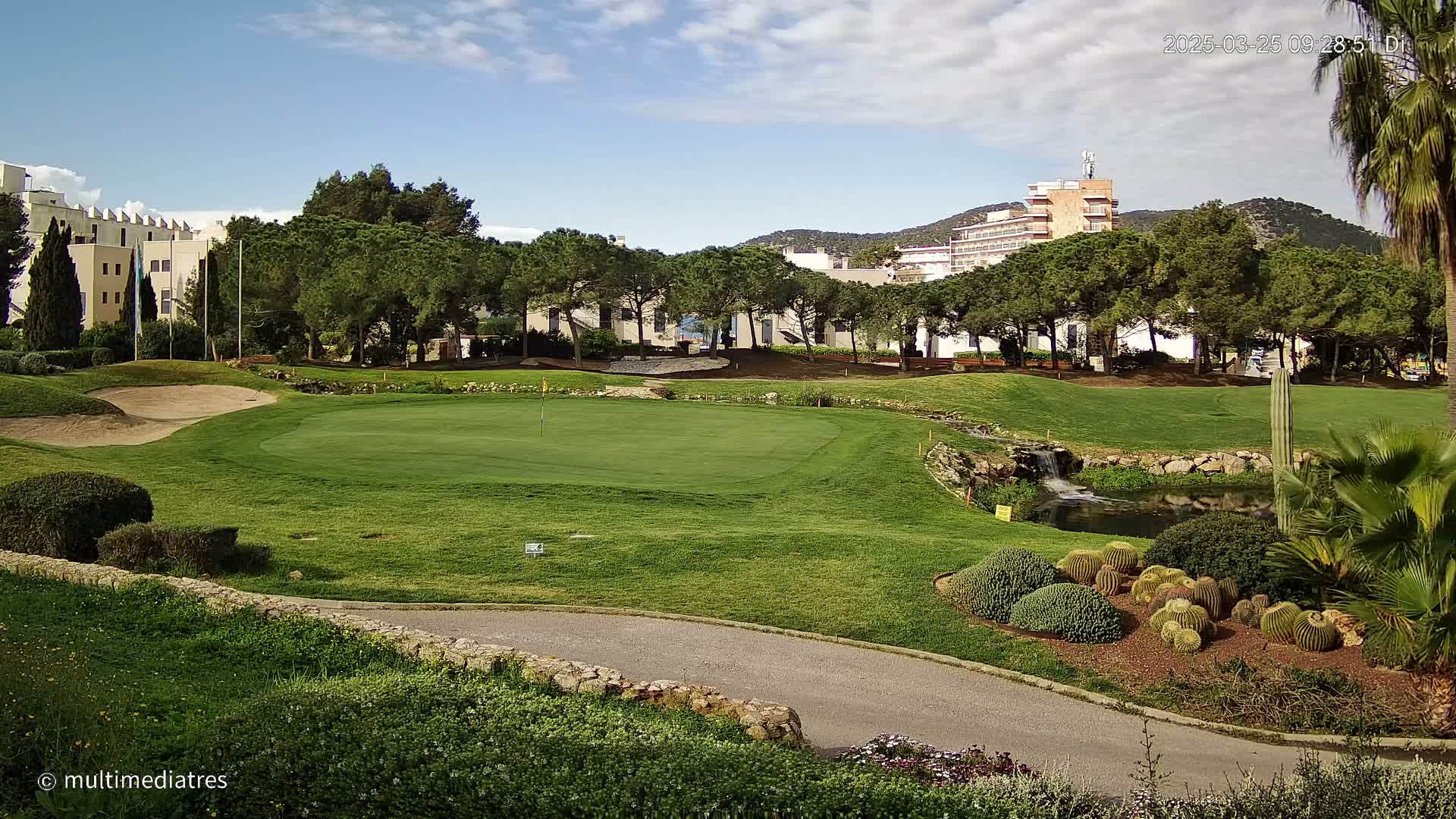 A golf course green with a sand trap and a small pond, situated near buildings and lush vegetation under a mostly clear sky.