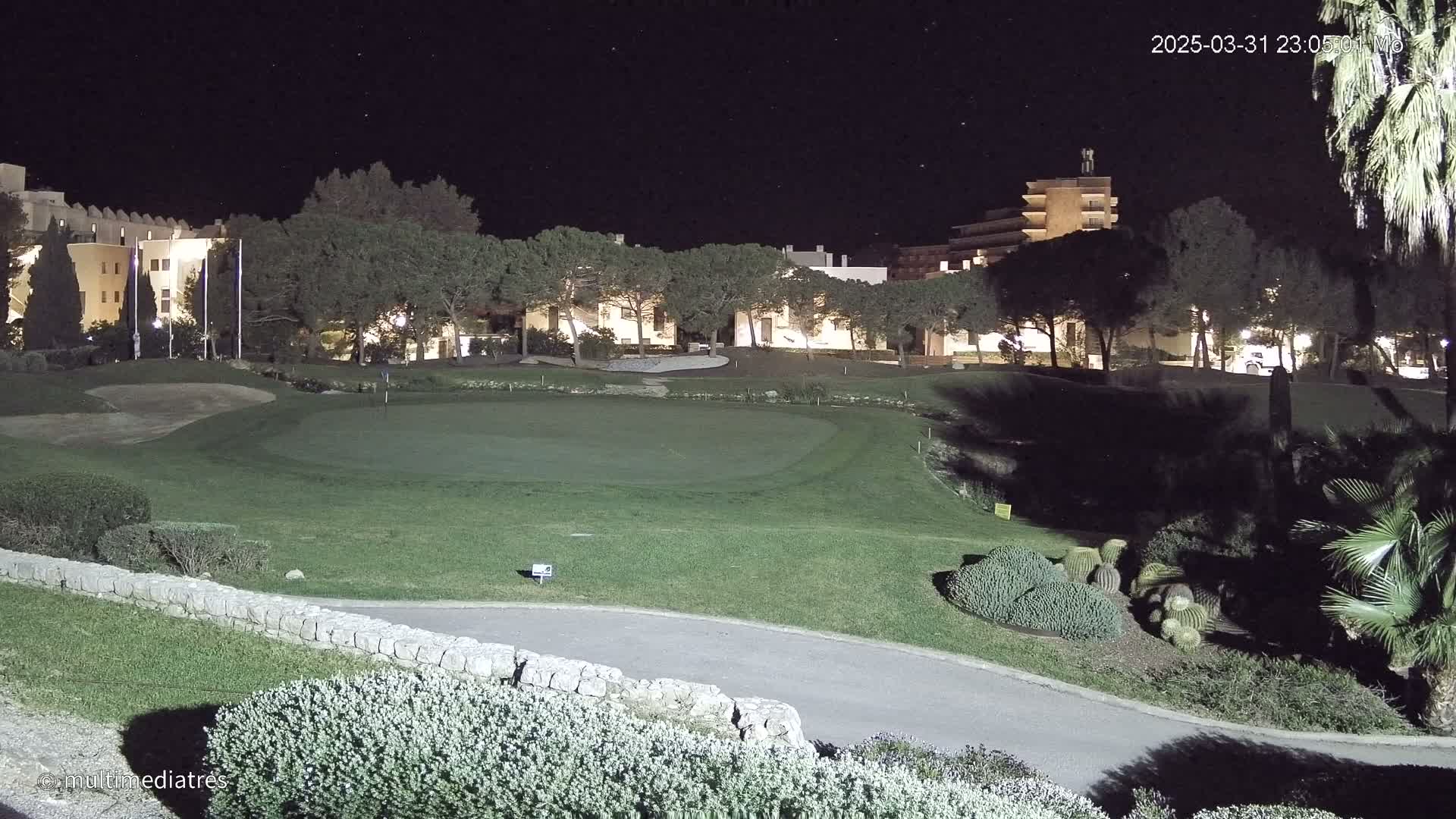A nighttime view of a golf course green illuminated by artificial lights, with buildings and trees in the background under a clear, starry sky.
