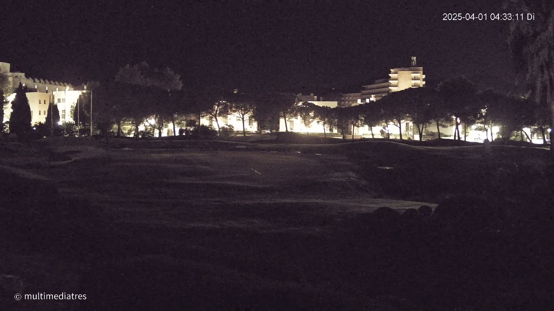 At night, illuminated buildings are visible behind a dark, uneven expanse of land that appears to be a grassy field or park, under a clear sky.
