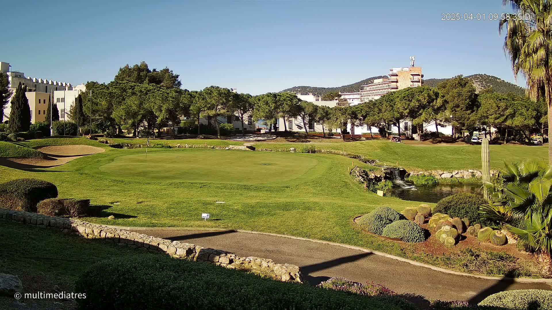 A golf course green with a small pond and waterfall is surrounded by lush green trees and buildings under a clear blue sky.