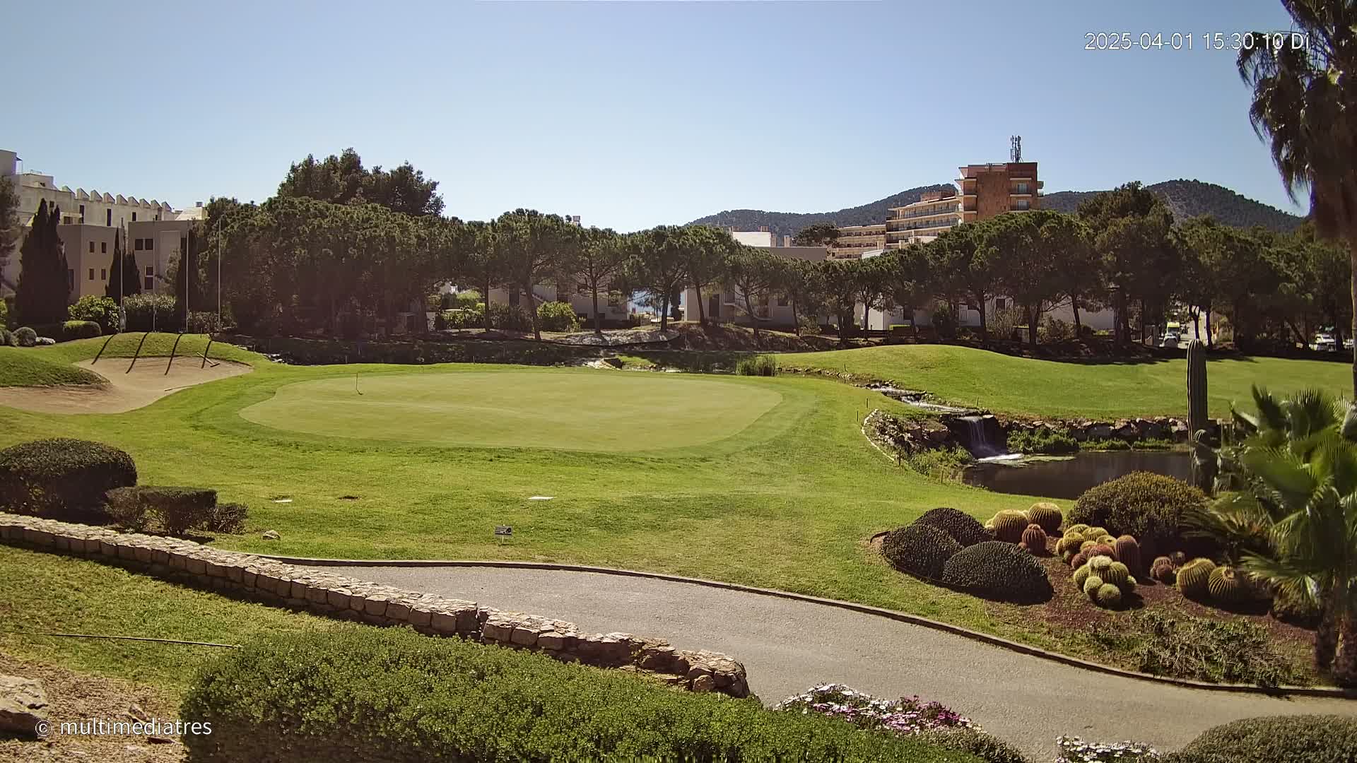 A sunny day reveals a putting green on a golf course, bordered by a stone wall and landscaping that includes succulents and a small pond with a waterfall, with buildings and trees in the background.