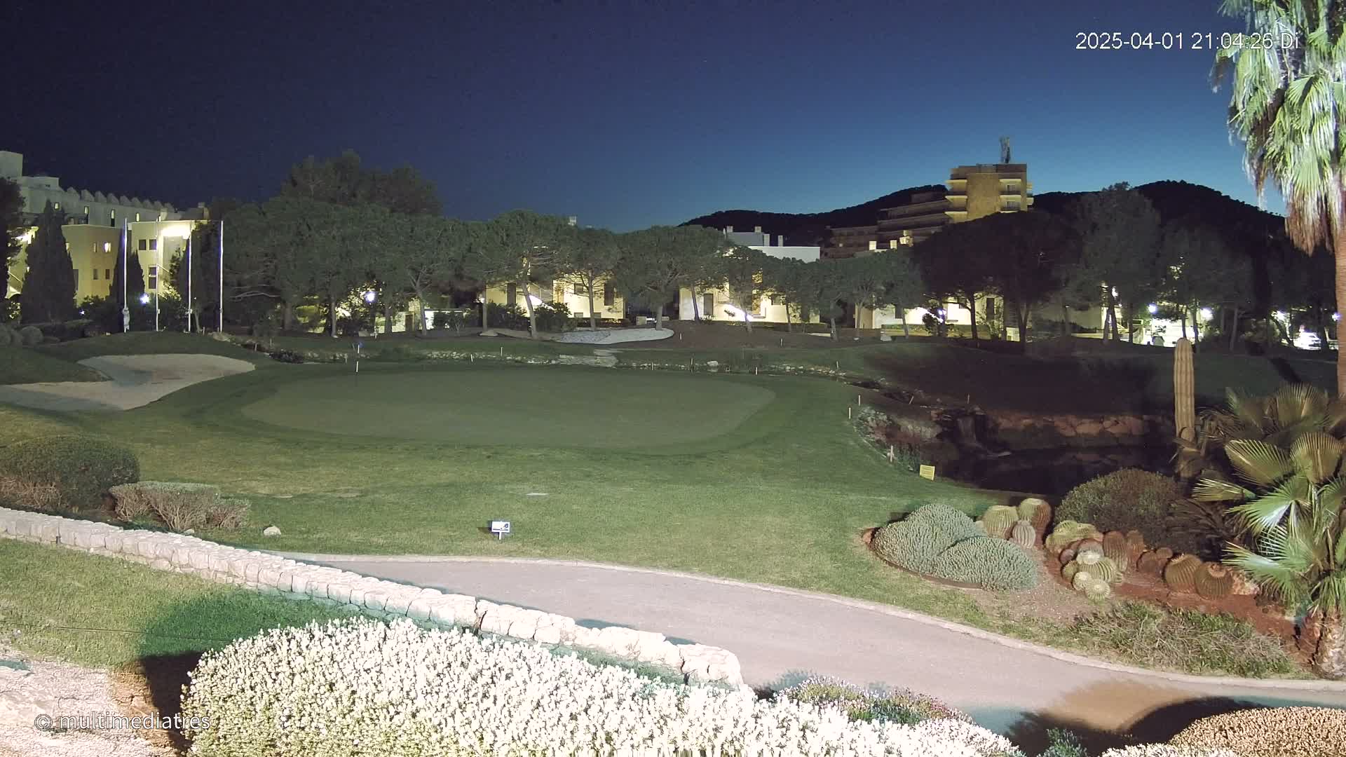 A putting green on a golf course at night, illuminated by artificial light, is situated in front of low-rise buildings and a hill under a dark blue, clear sky.