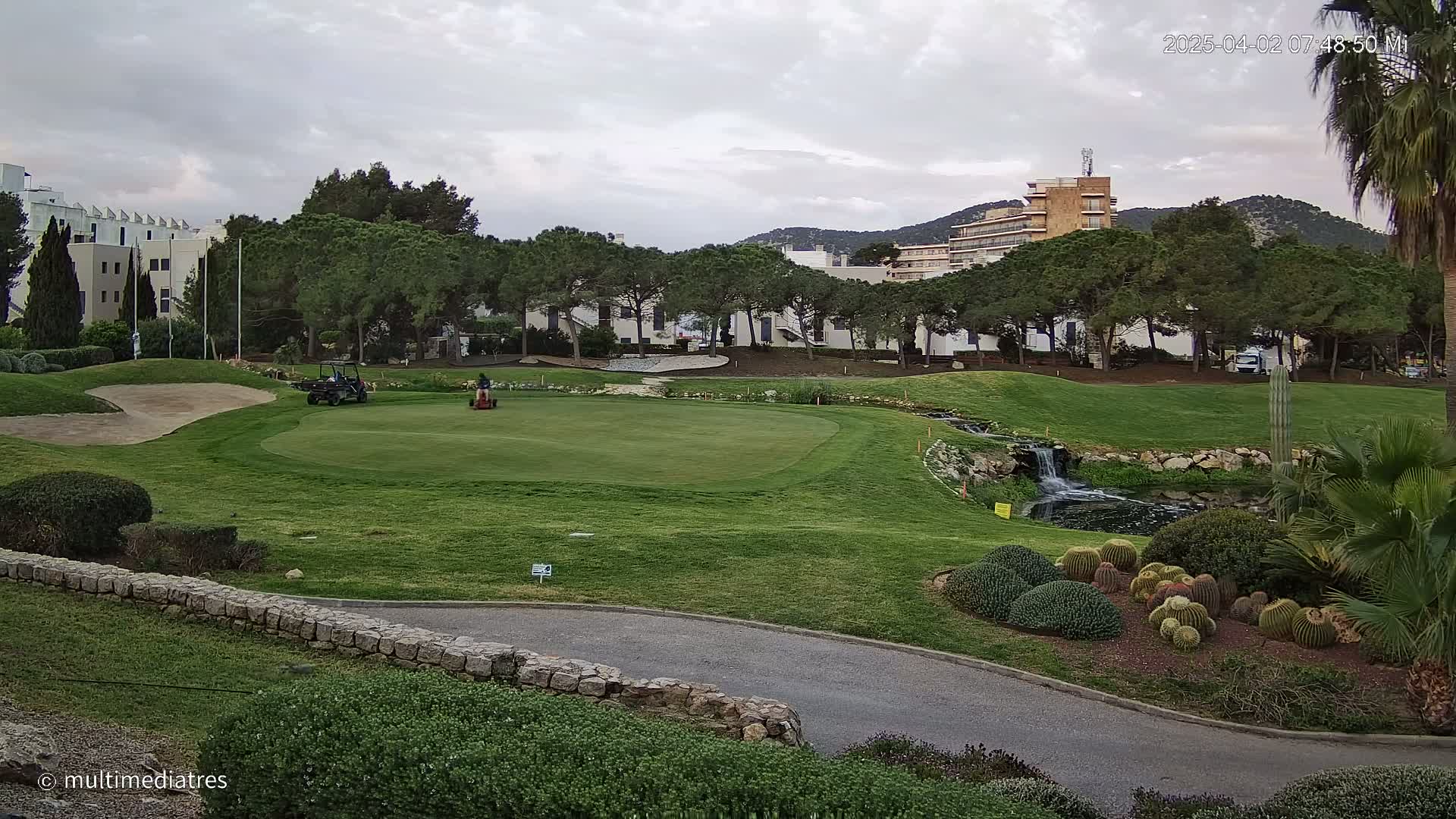 A person operates a riding lawnmower on a green golf course with a small waterfall and cacti in the foreground, under a cloudy sky.