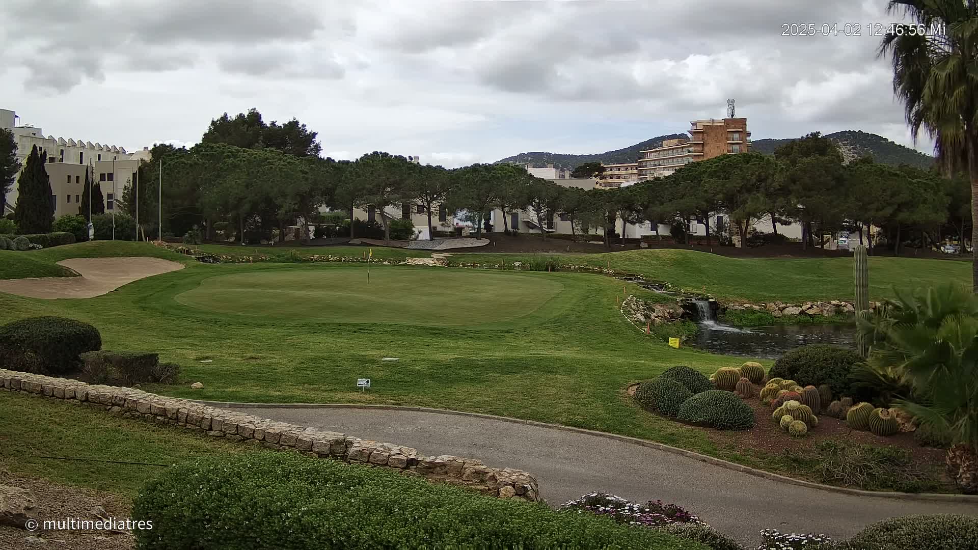 A golf course green with a small pond and waterfall, under a cloudy sky, is situated near buildings and trees.