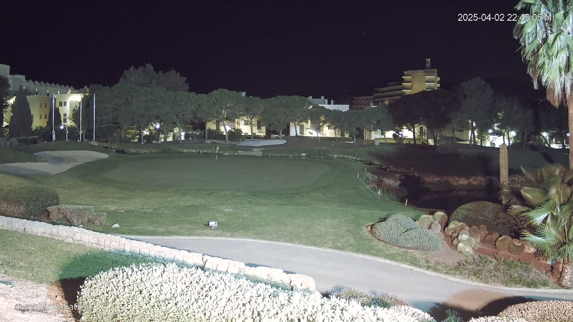 A nighttime view of a golf course putting green, bordered by low stone walls and landscaping, with buildings and trees illuminated in the background.