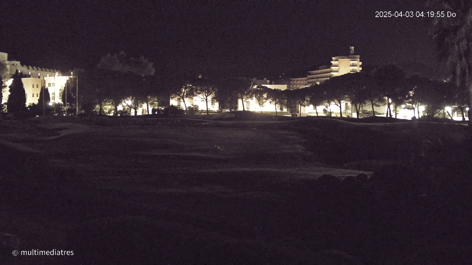 At night, a line of illuminated buildings is partially obscured by a row of dark trees, beyond which is a low, dark, uneven expanse of land.