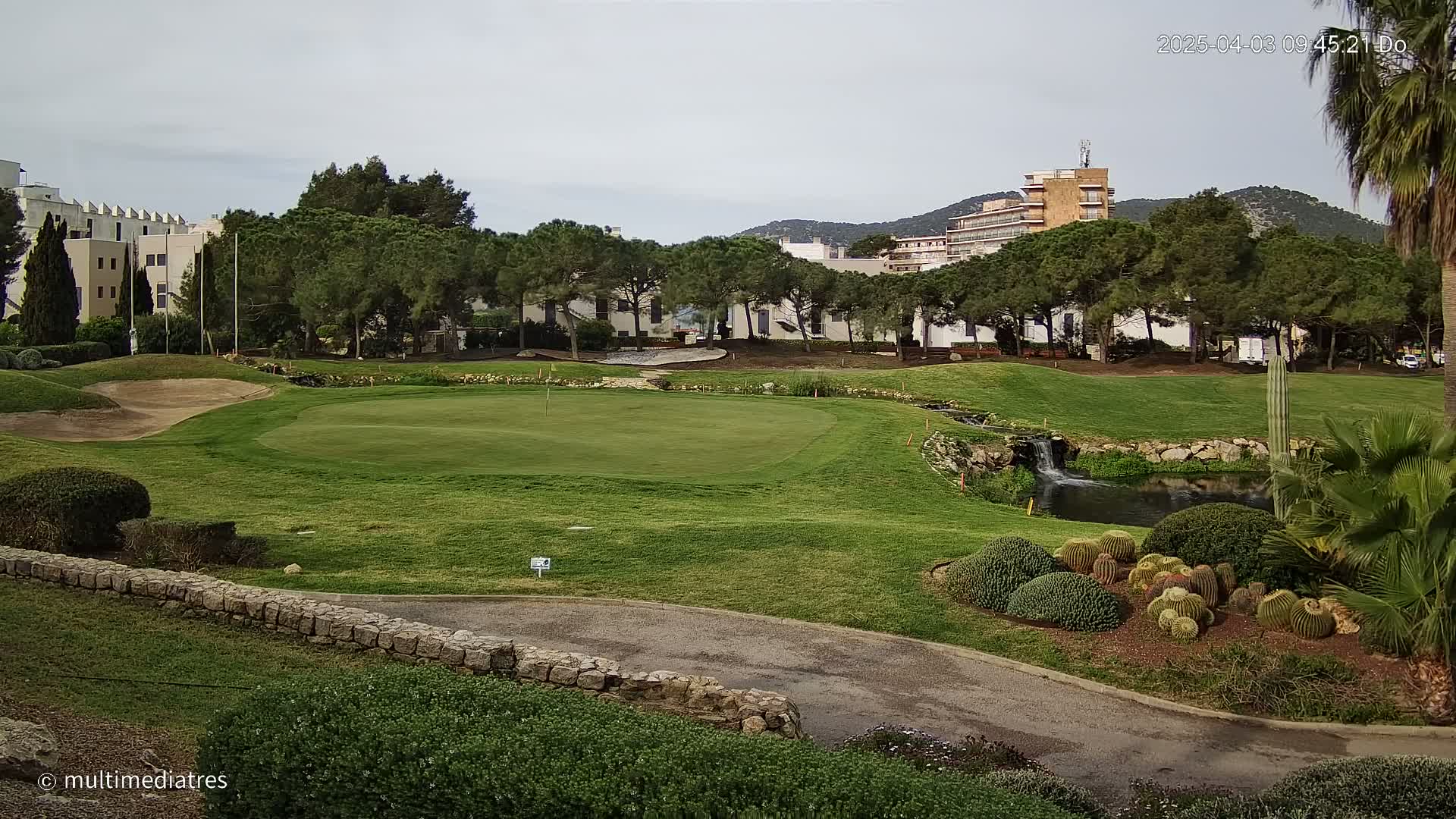 A golf course green with a small pond and several buildings in the background under an overcast sky.