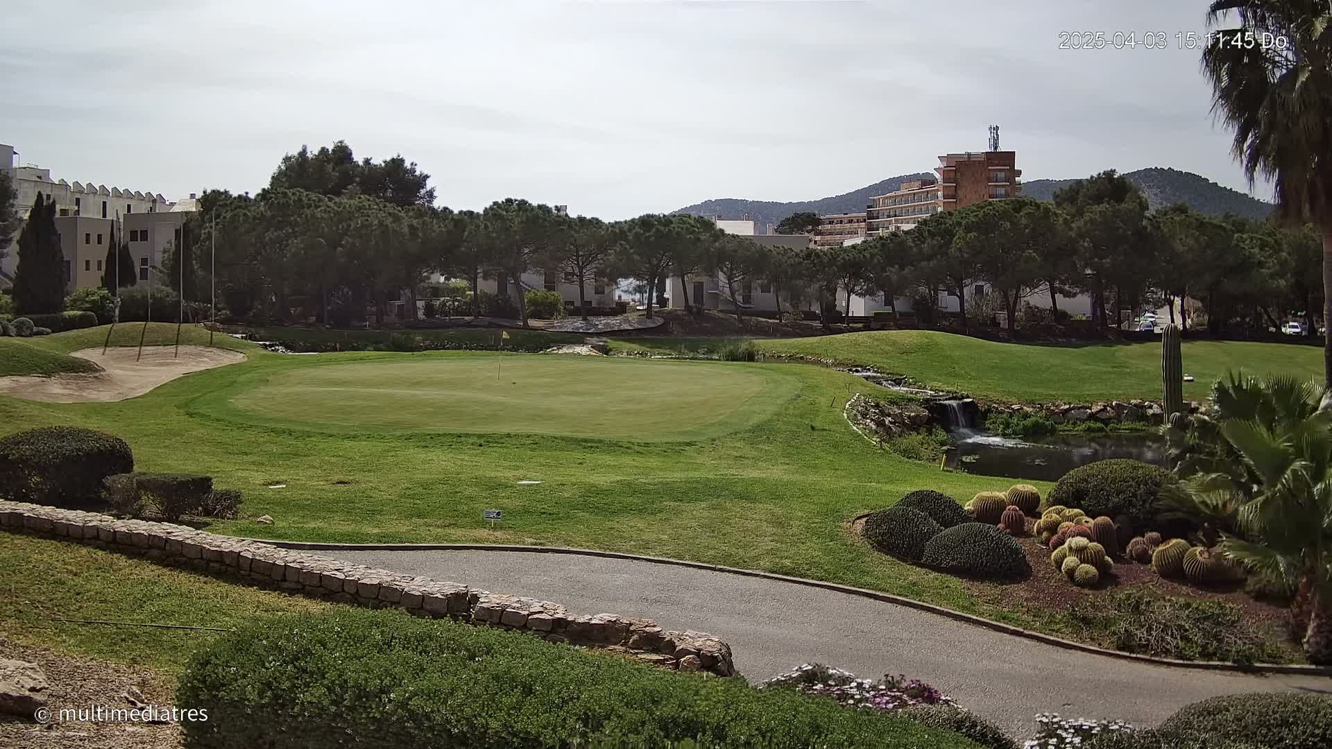 A golf course putting green with a small pond and surrounding landscaping, situated near buildings under a partly cloudy sky.