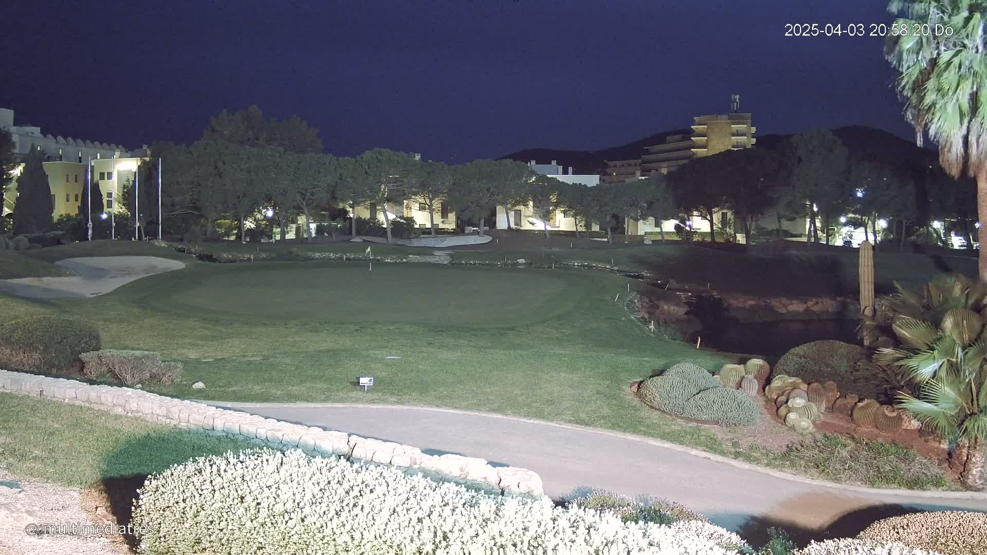 A well-lit putting green at night is bordered by landscaping, a low stone wall, and buildings in the background under a dark sky.