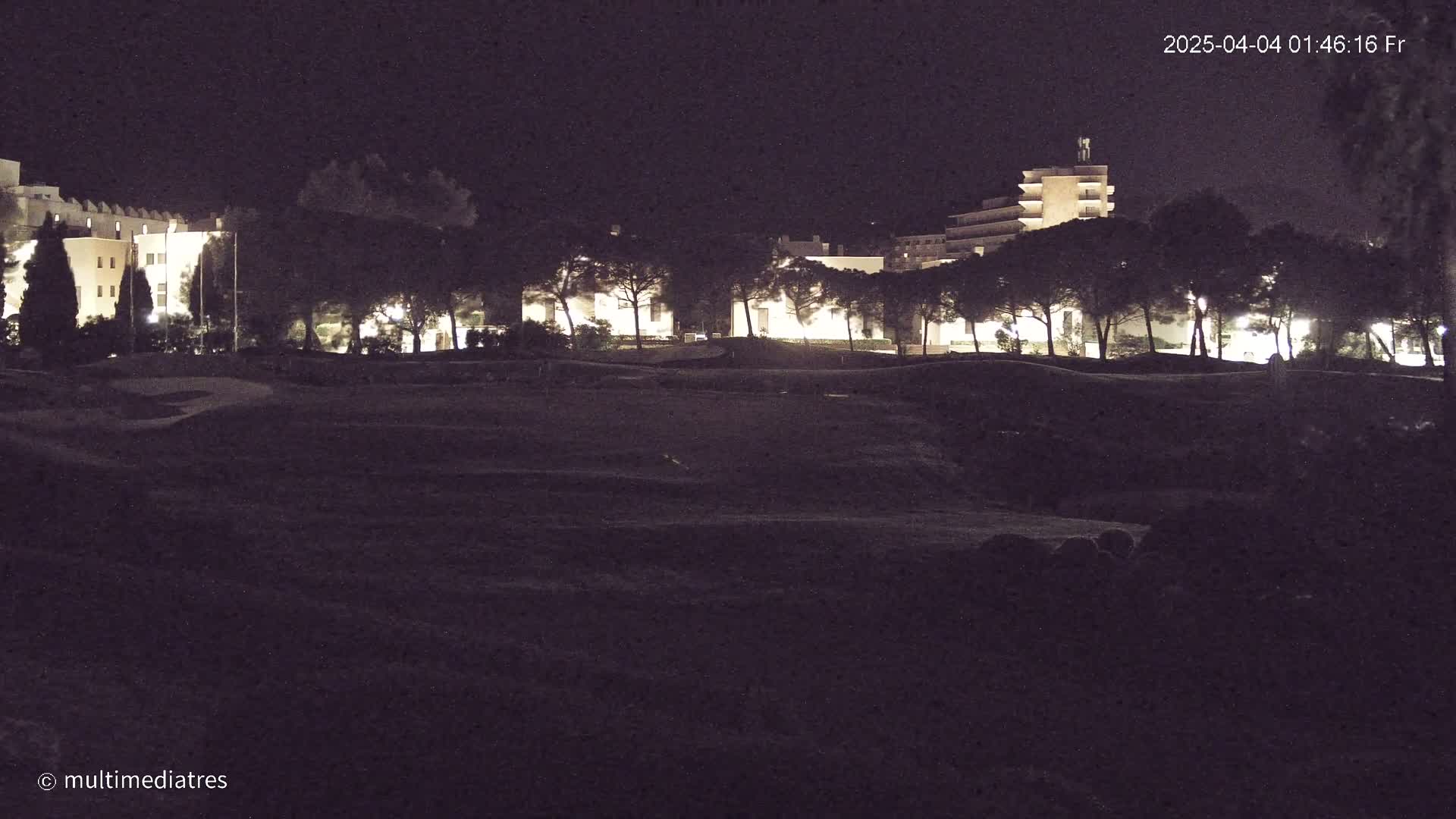 At night, illuminated buildings are visible beyond a dark, uneven field of land covered with short vegetation, lined with a row of dark trees.