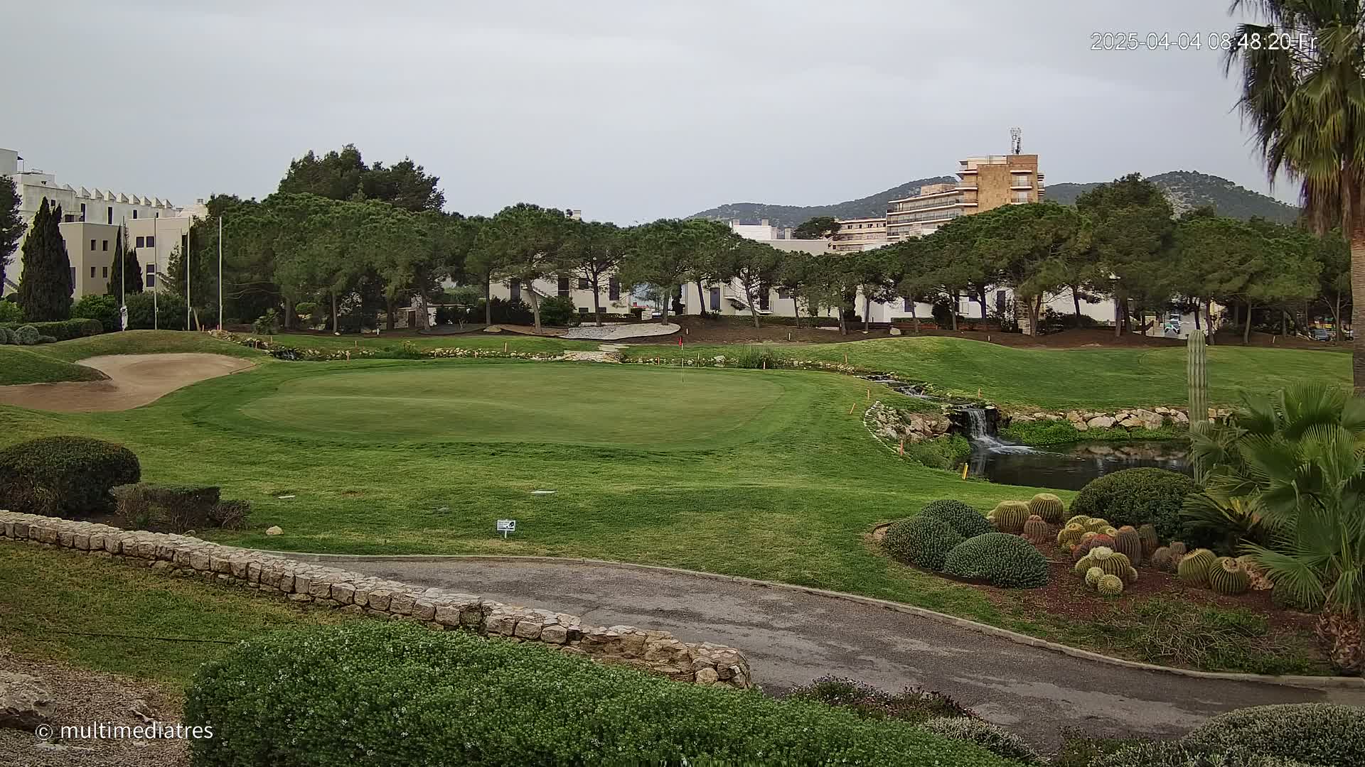 A golf course putting green, bordered by a stone wall and landscaping that includes a small pond and cacti, sits under an overcast sky near buildings and trees.