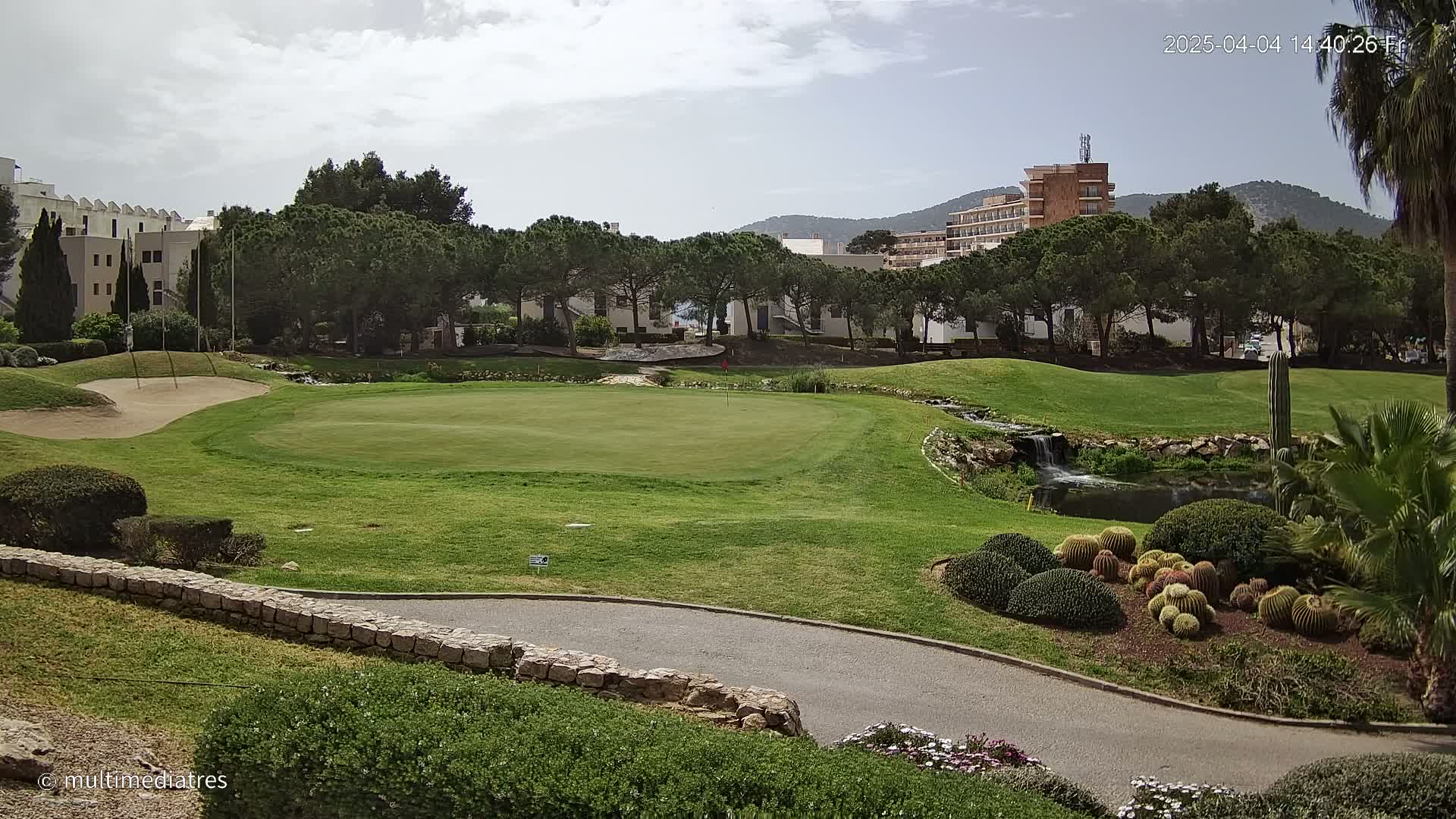 A golf course green with a sand trap, a small waterfall, and numerous cacti and succulents, is situated next to a row of buildings under a partly cloudy sky.