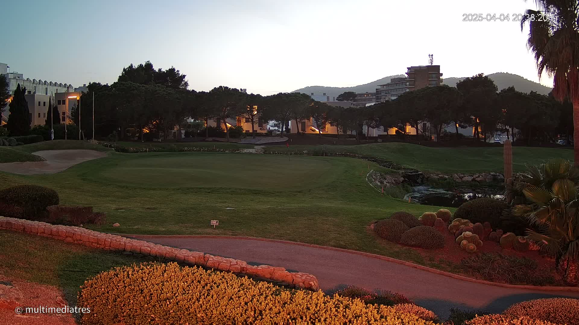 A golf course green at twilight, with buildings and trees in the background under a clear sky.
