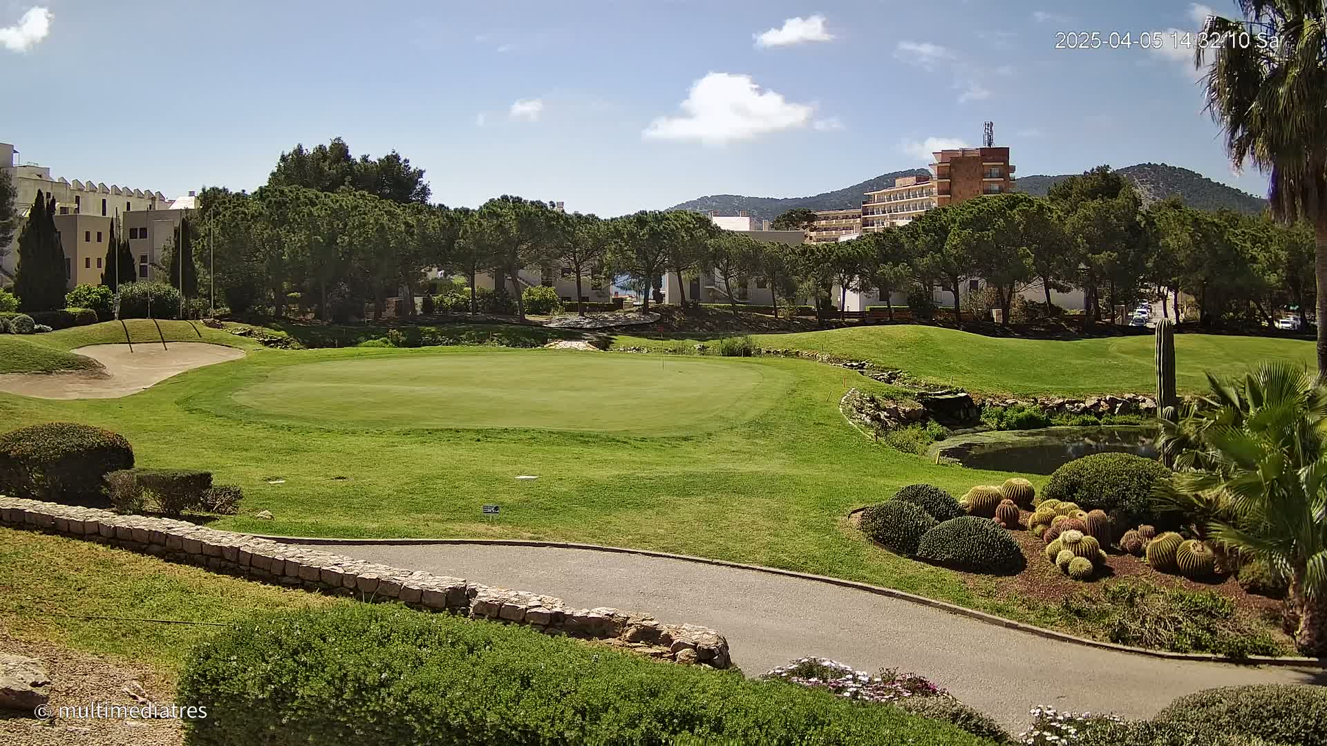 A golf course putting green with sand traps and landscaping is situated near buildings and trees under a sunny sky.