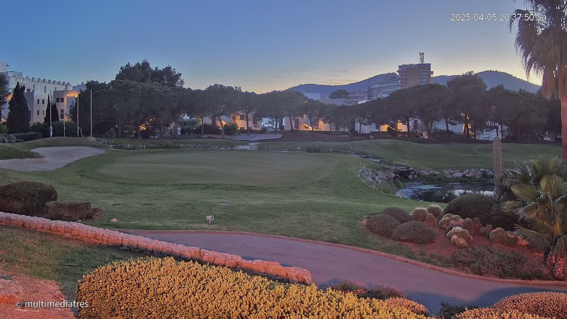 A golf course at dusk, with buildings and trees in the background, under a clear twilight sky.