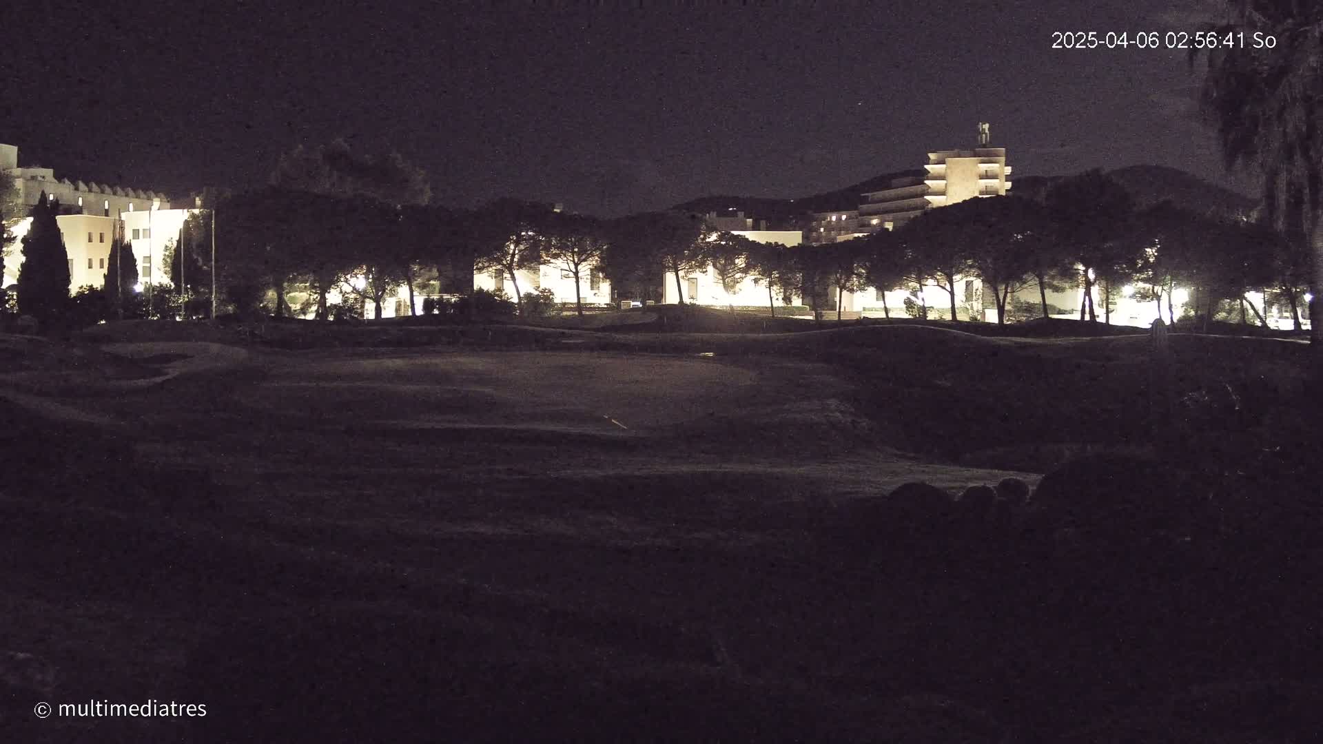 At night, a dark, undulating grassy area lies in front of a line of illuminated buildings and trees.