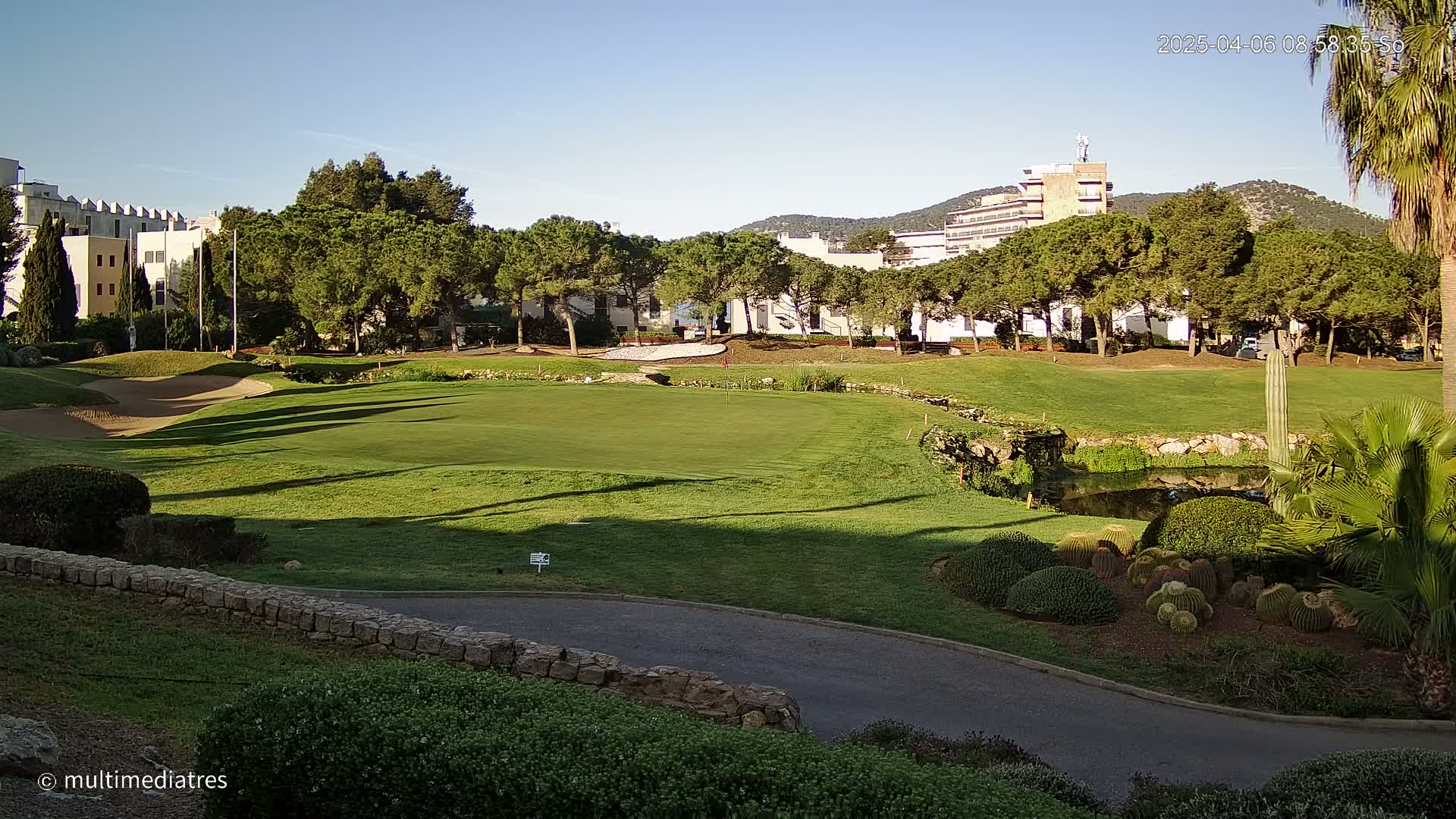 A sunny golf course with green grass, sand traps, a small pond, and buildings in the background.