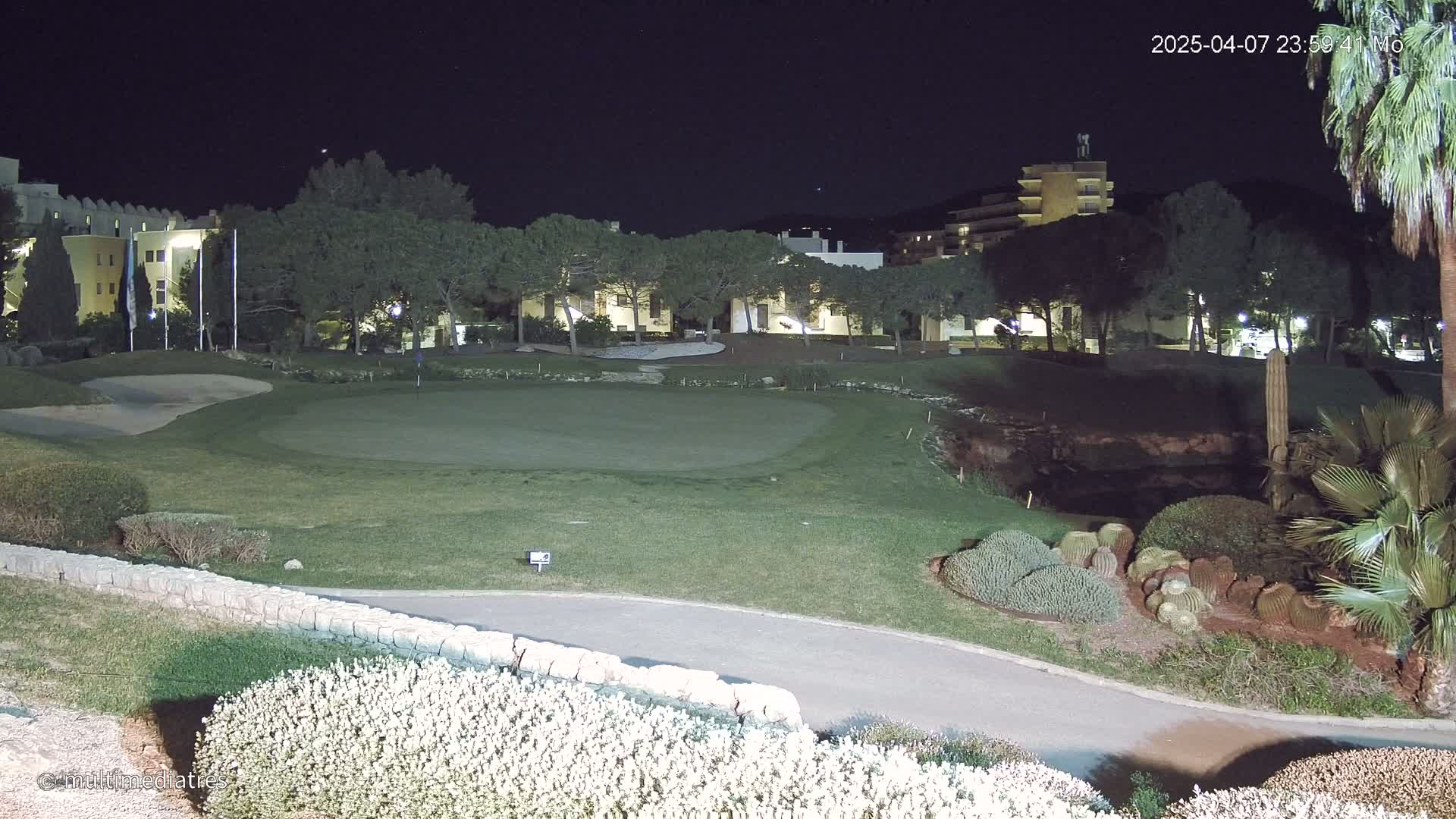 A brightly lit golf course green at night, bordered by trees, shrubs, and buildings.
