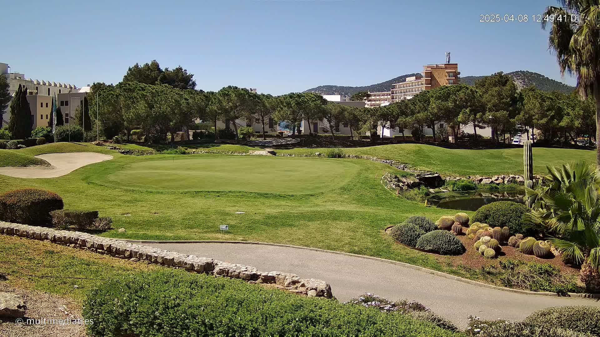 A golf course putting green is nestled among lush green landscaping, a small pond, and buildings in the background under a clear, sunny sky.