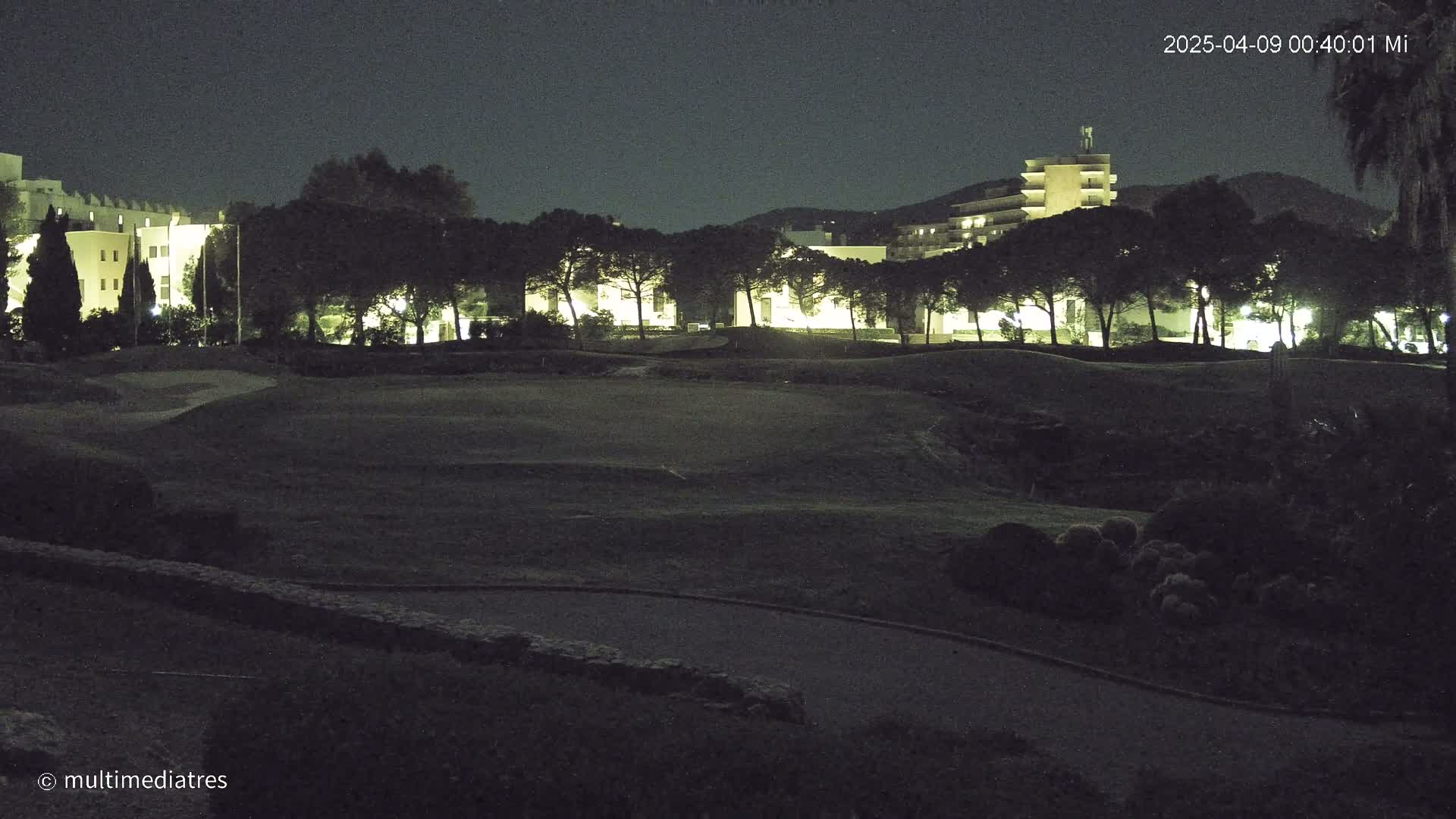 A nighttime view of a golf course with illuminated buildings and trees in the background under a dark sky.