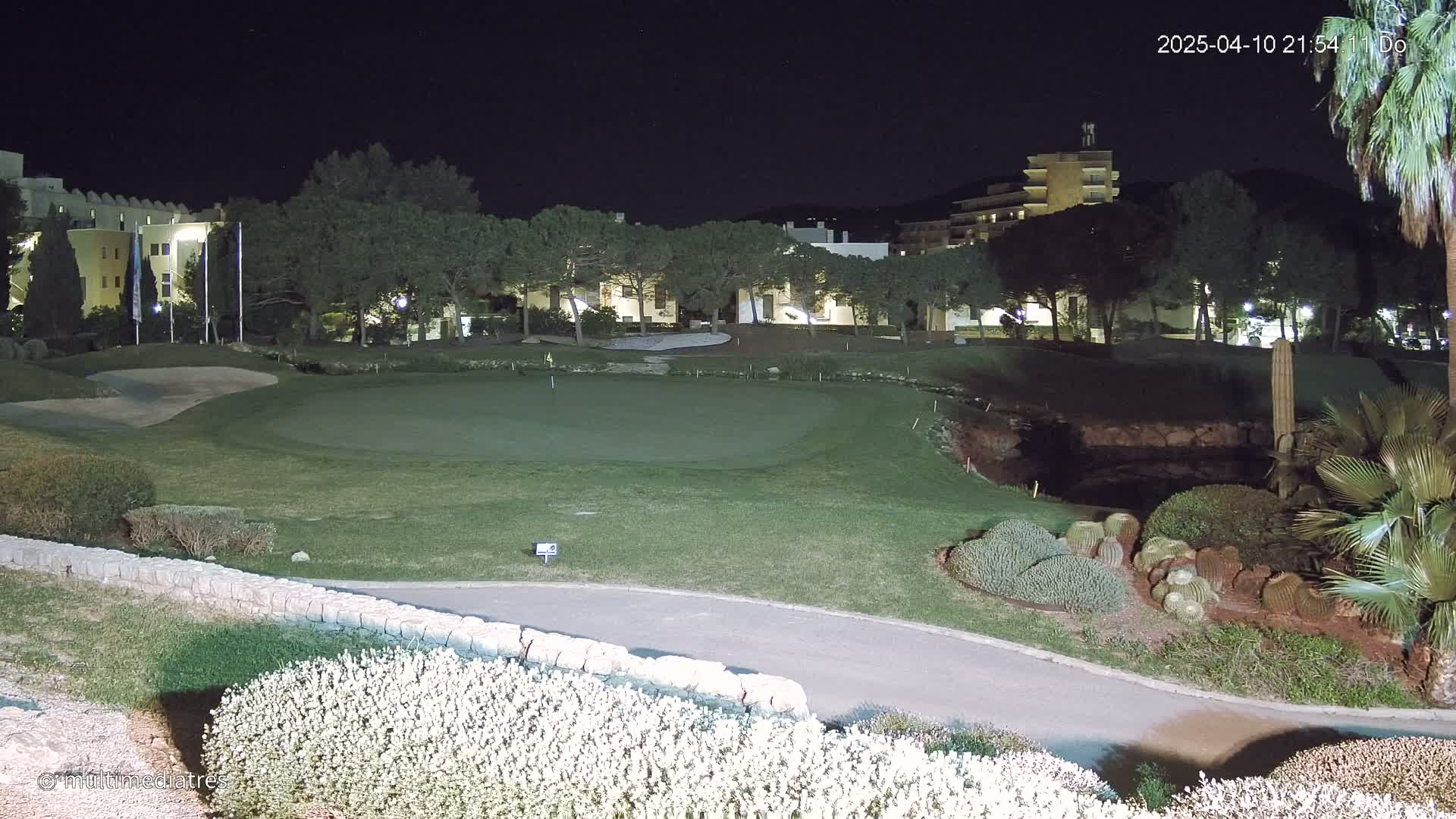 A nighttime view of a putting green on a golf course, illuminated by artificial lights, with buildings and trees in the background.