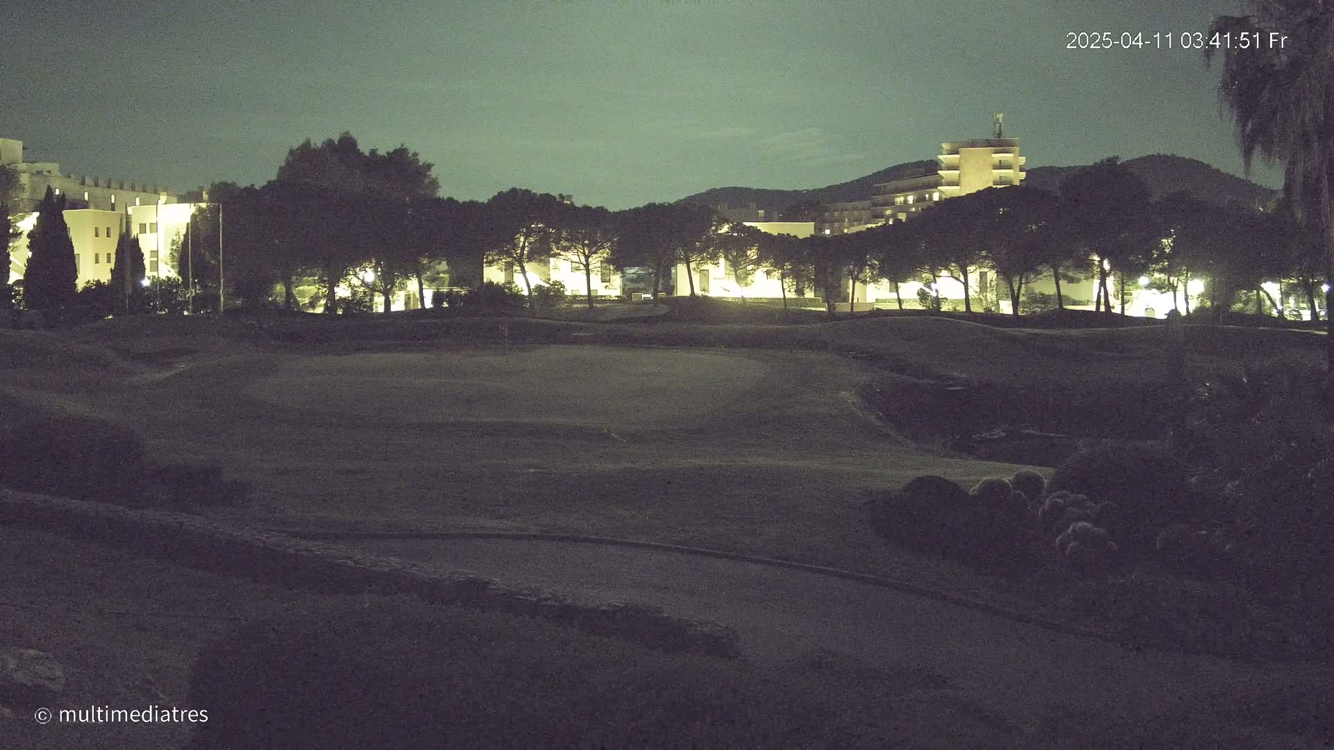 A nighttime view of a golf course with buildings and trees illuminated in the background under a dark sky.