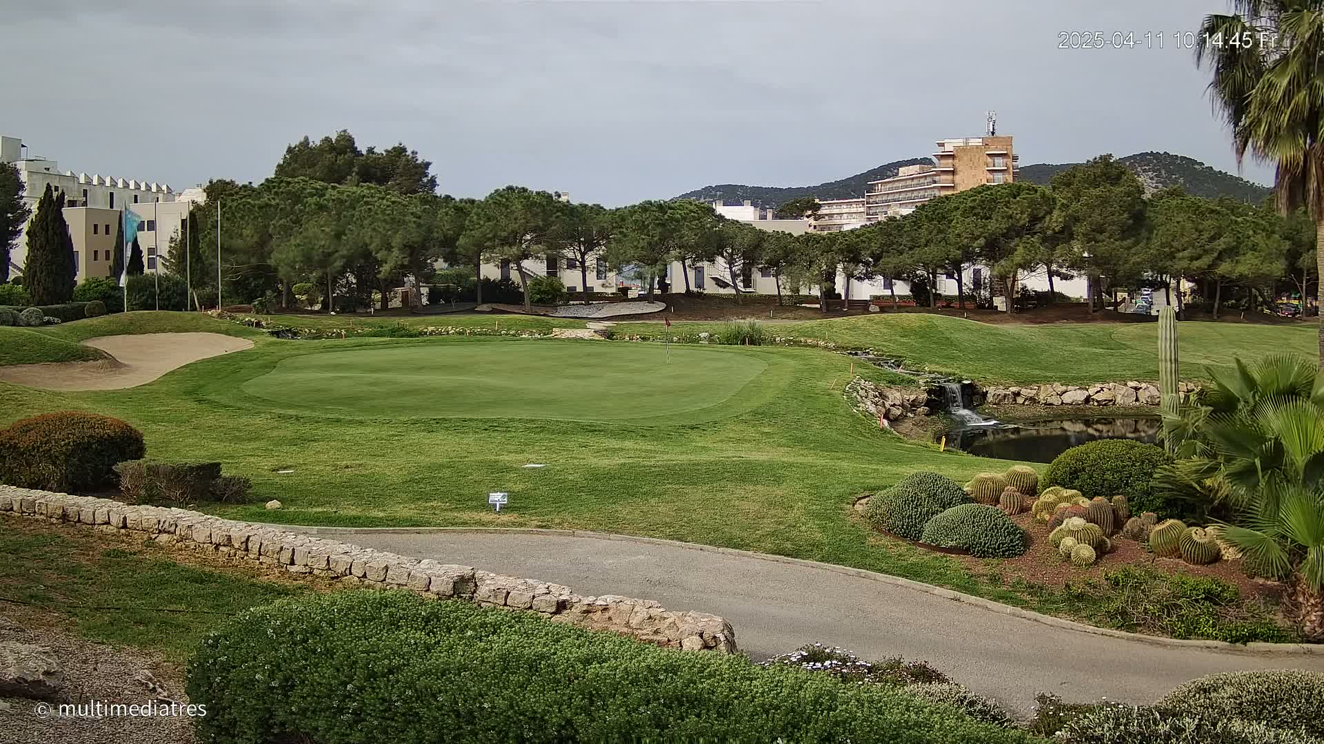 A golf course green with a small pond and waterfall, surrounded by lush landscaping and buildings in the background under a cloudy sky.