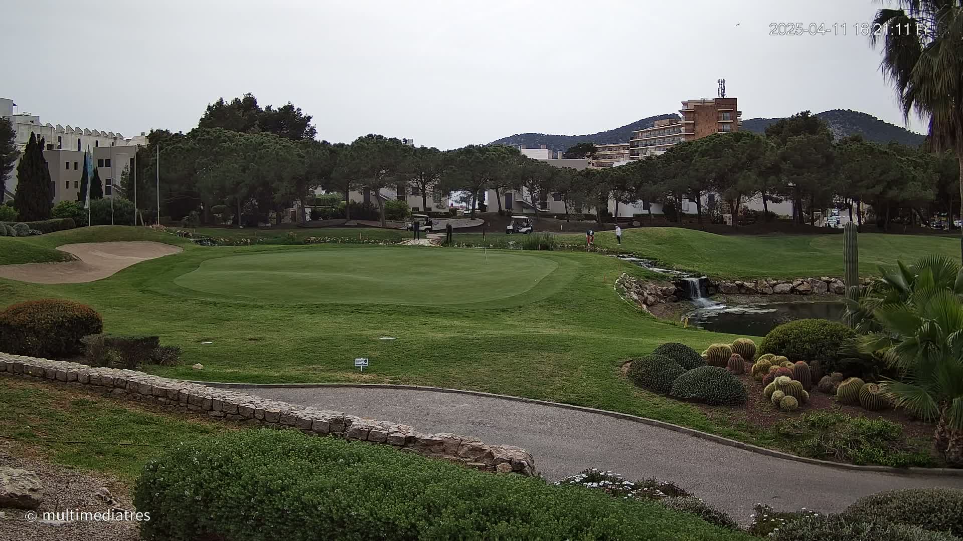 A golf course with a green, sand trap, small pond, and several golfers is visible, under an overcast sky.