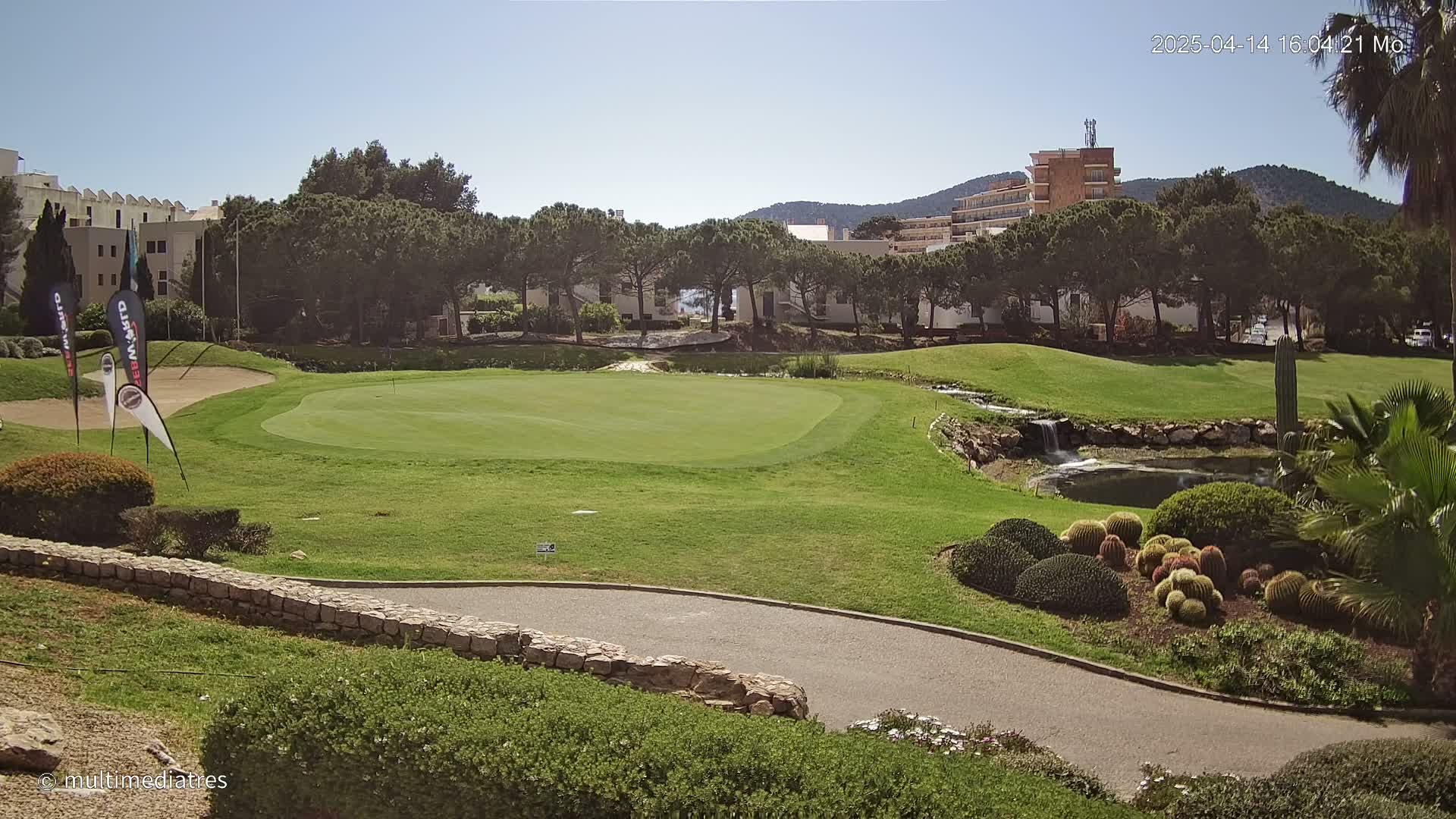 A golf course green with a small pond and waterfall, surrounded by trees and buildings under a sunny sky.